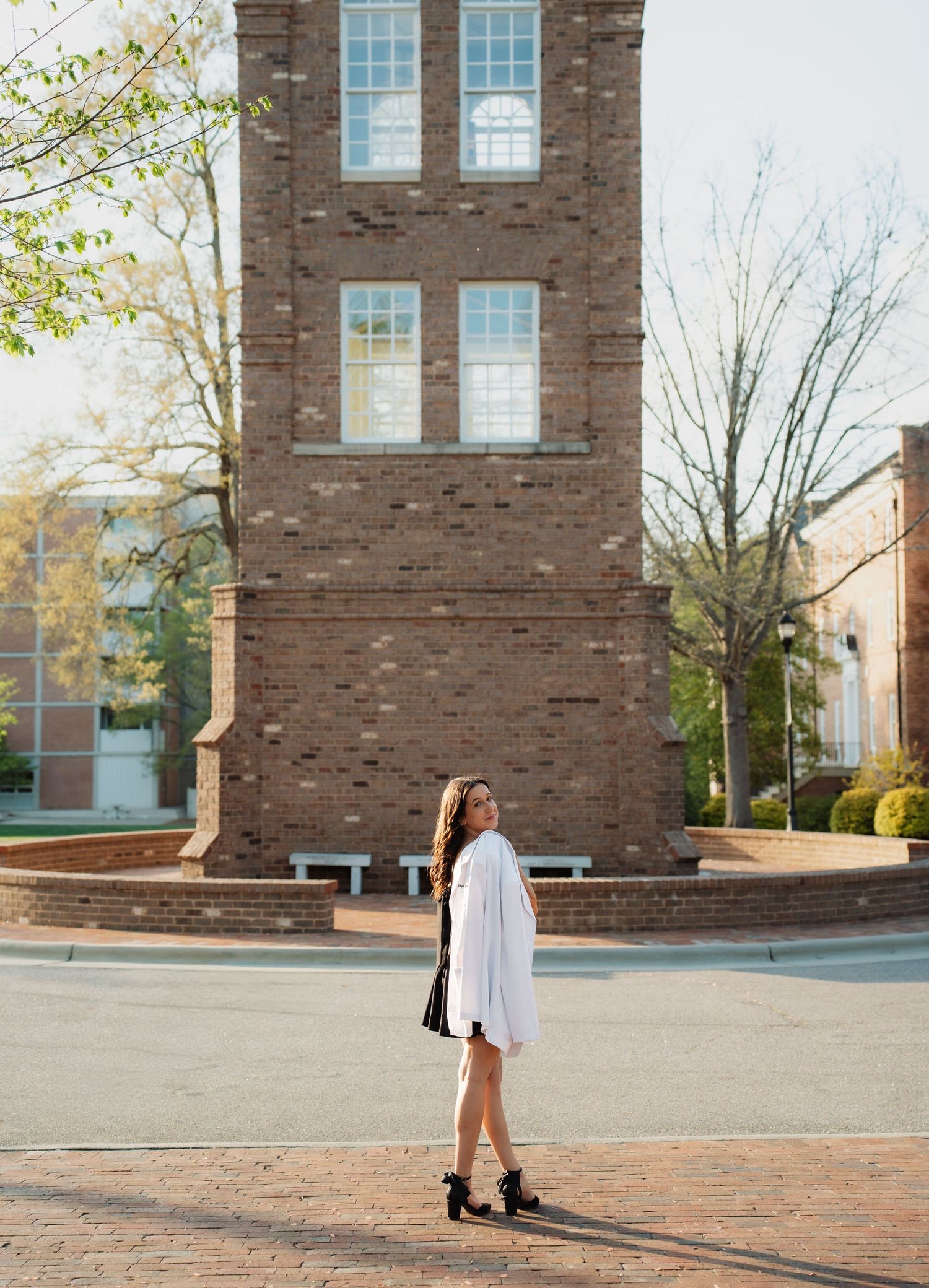Young woman in white dress and heels poses on brick plaza beside historic red brick tower building on sunny day for her Graduation photos.