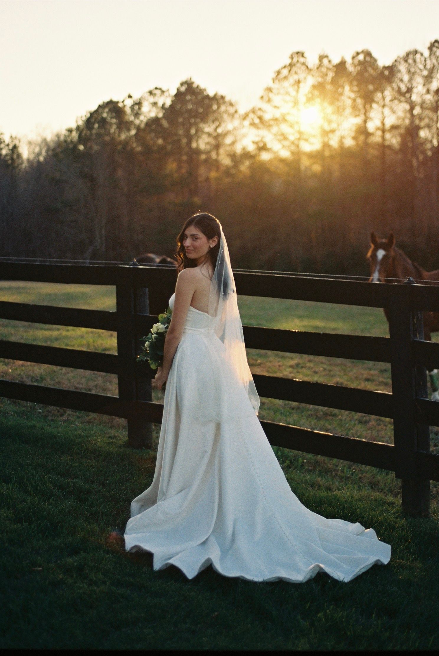 A bride in a flowing white wedding gown poses at sunset near a rustic wooden fence in a pastoral setting.