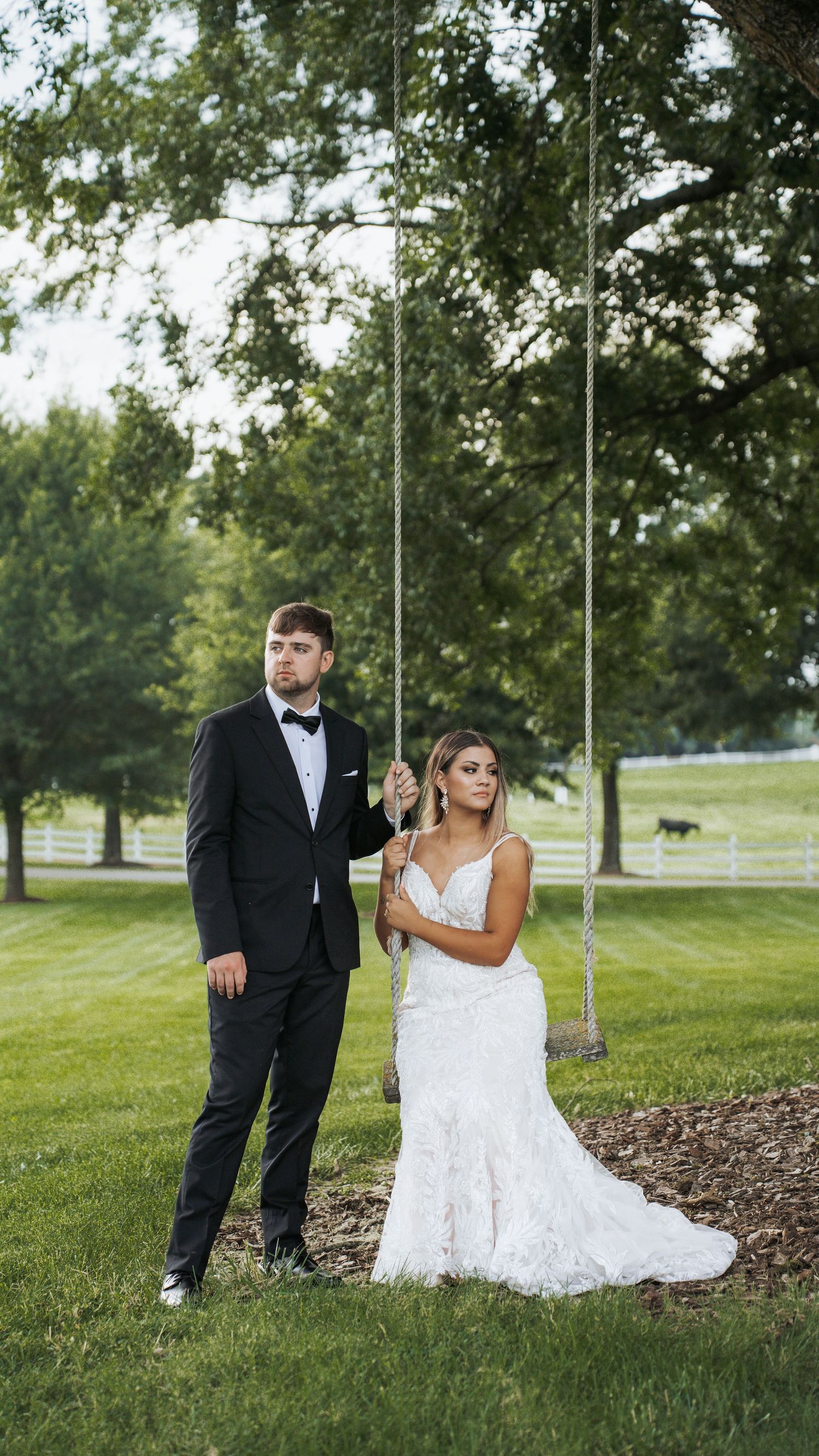 A couple poses playfully on a grassy hill under tall trees.