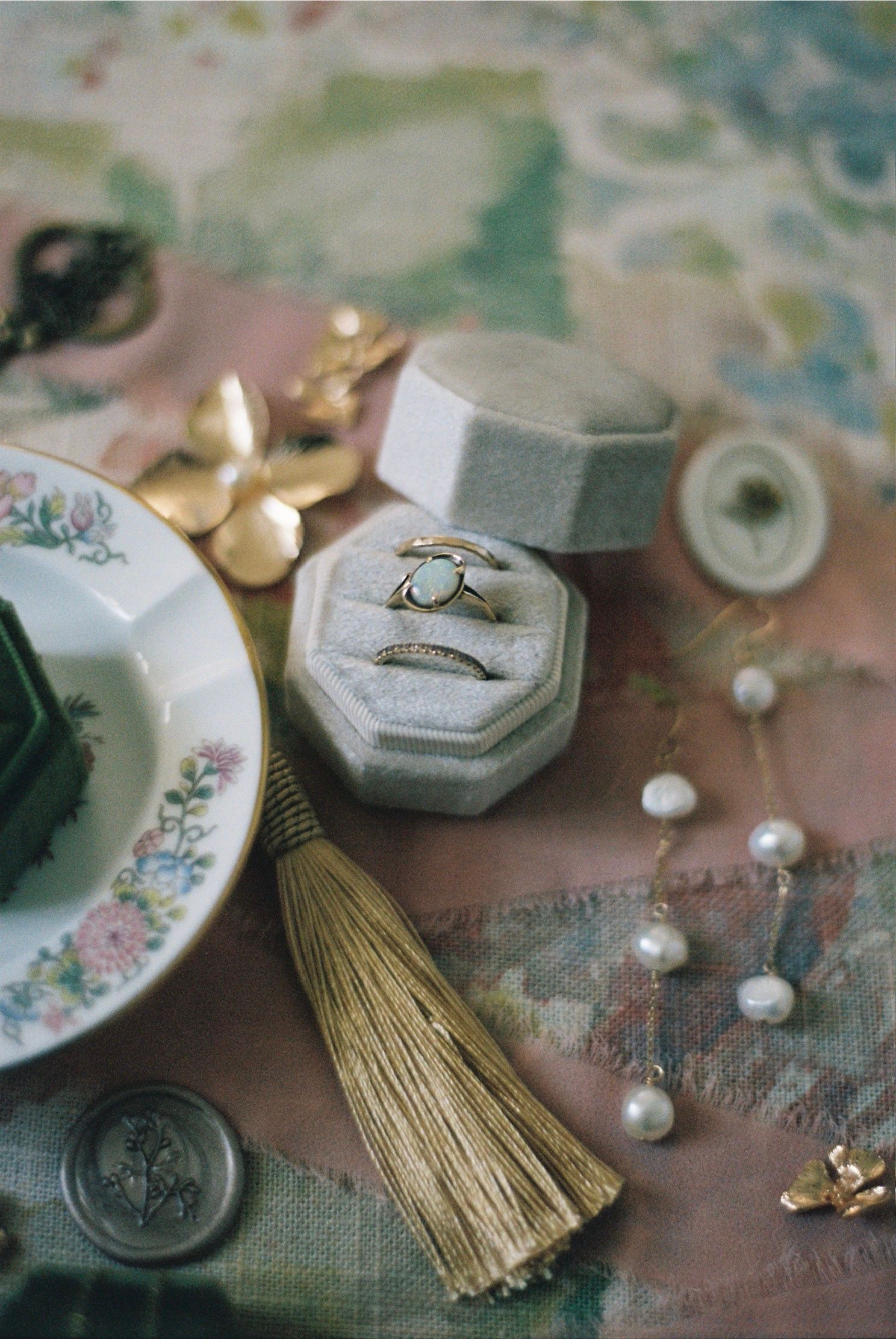 Vintage-inspired wedding details with pearls, gold tassels, and floral plates arranged on a dusty pink textured surface.