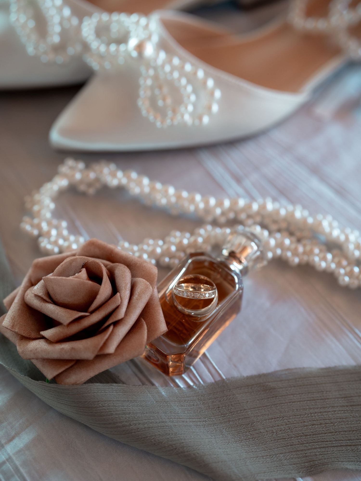 Wedding accessories including a dusty rose flower, pearl necklace, perfume bottle and white bridal shoes arranged elegantly.