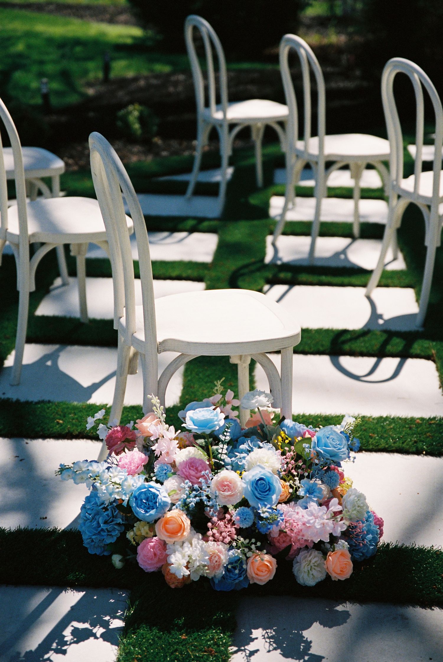 White garden chairs arranged outdoors with a beautiful pink and blue floral arrangement on the grass.