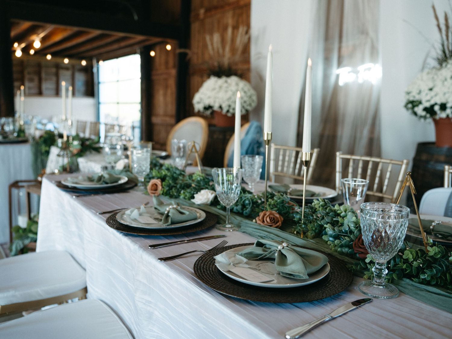 Elegant table settings with greenery garlands and crystal glasses in a rustic barn venue for a wedding reception.