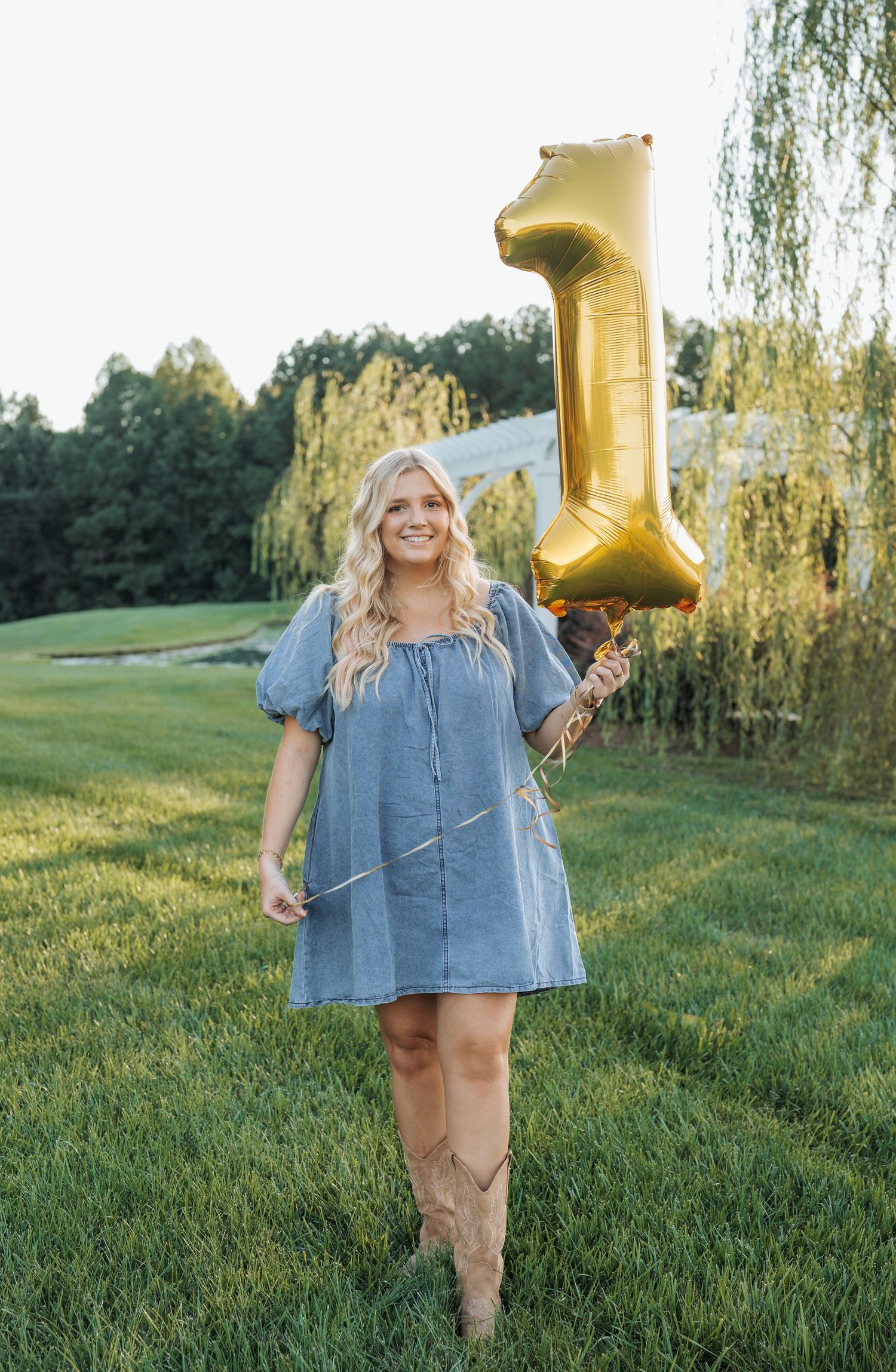 Person in blue dress holding gold number balloon in sunny garden setting.