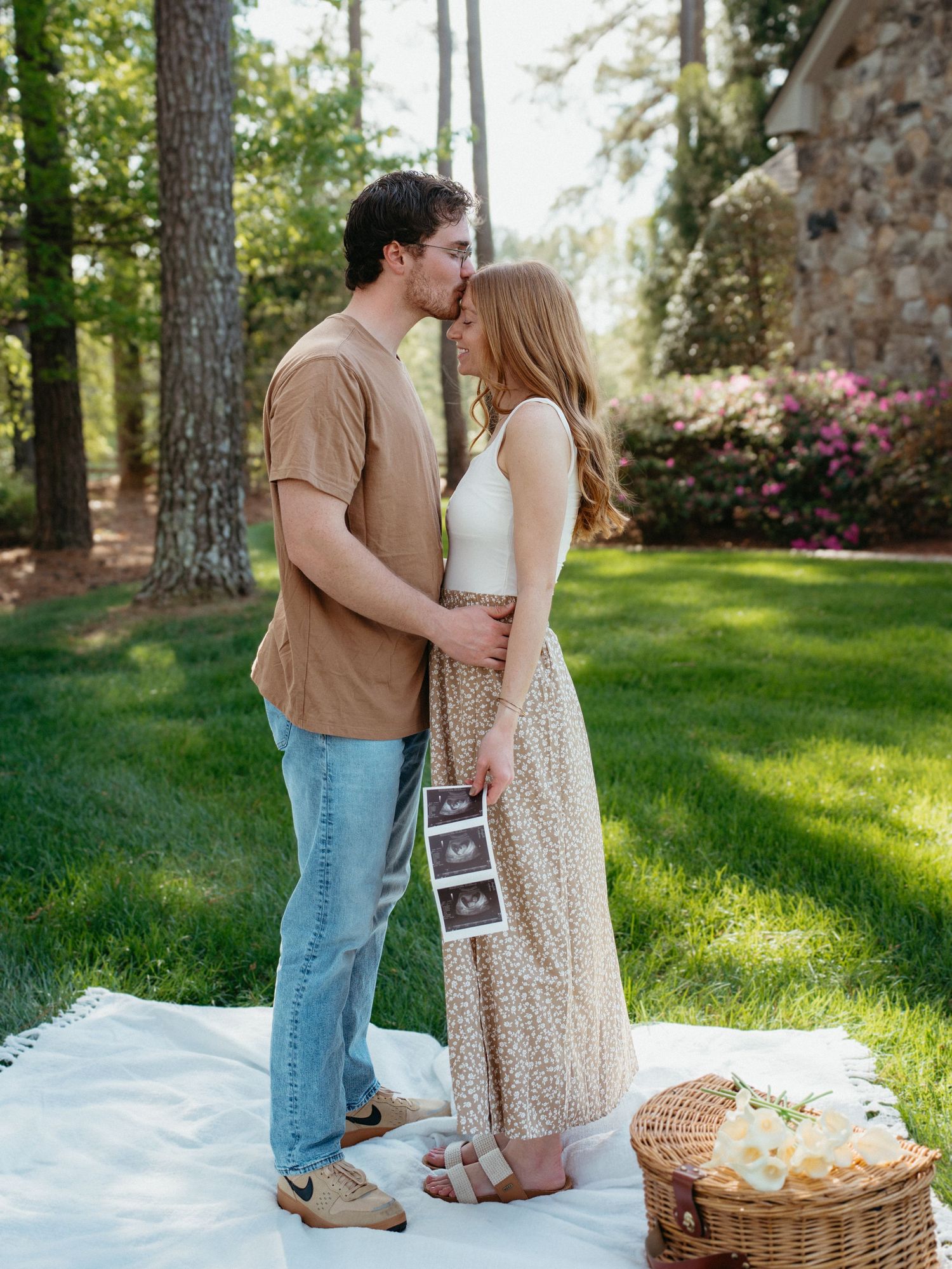 Couple kissing outdoors on white blanket holding ultrasound photos, pregnancy announcement in garden setting.