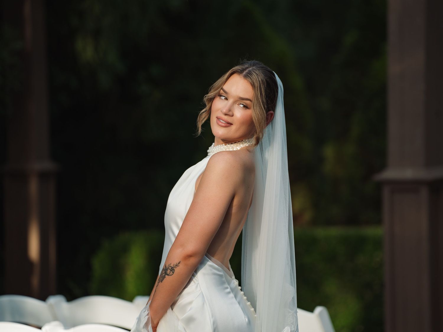 Bride in backless white gown and veil smiles over shoulder at outdoor wedding venue with lush greenery.