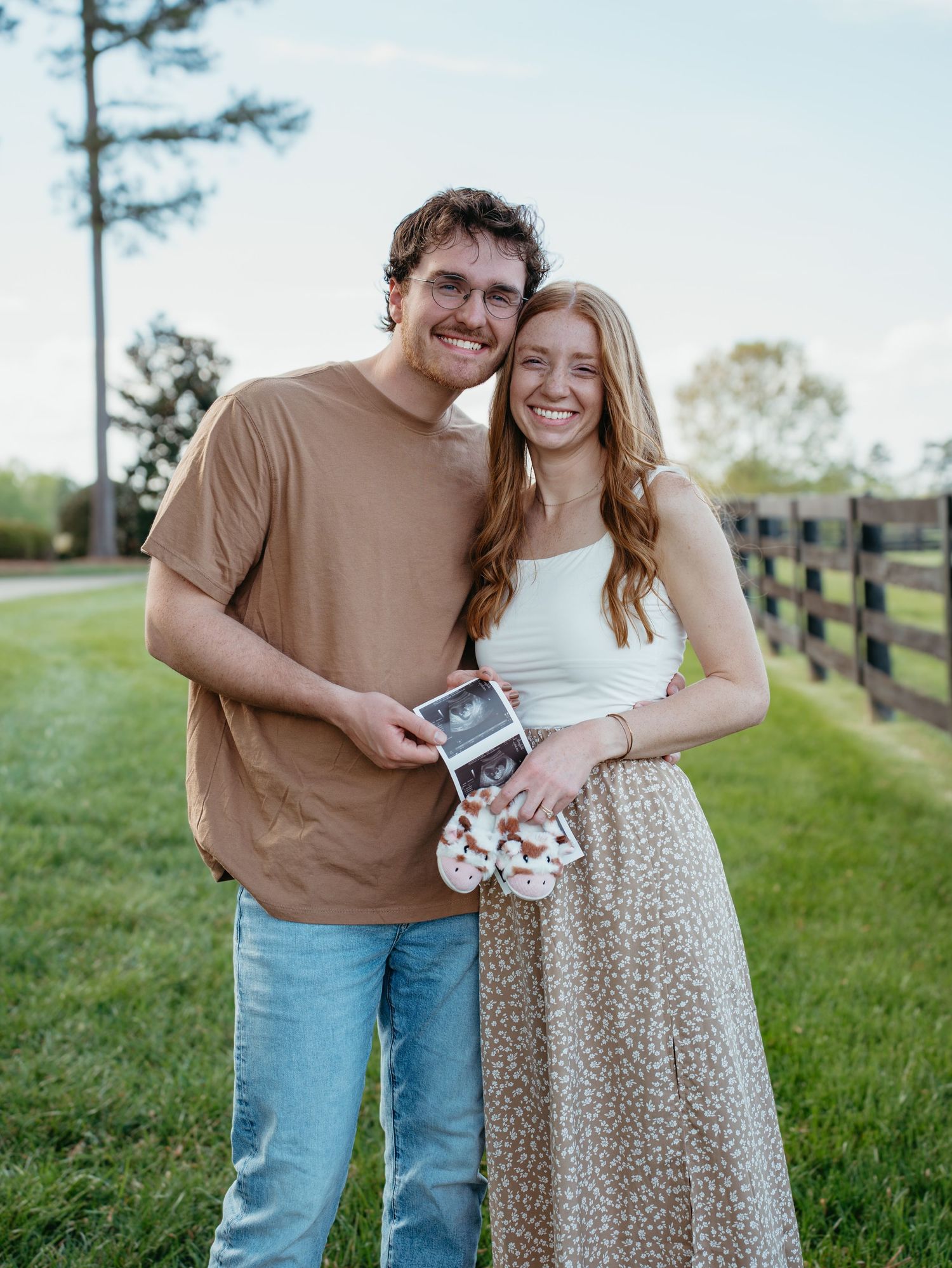 Happy couple smiling outdoors holding ultrasound photos and baby shoes, announcing pregnancy near a wooden fence.