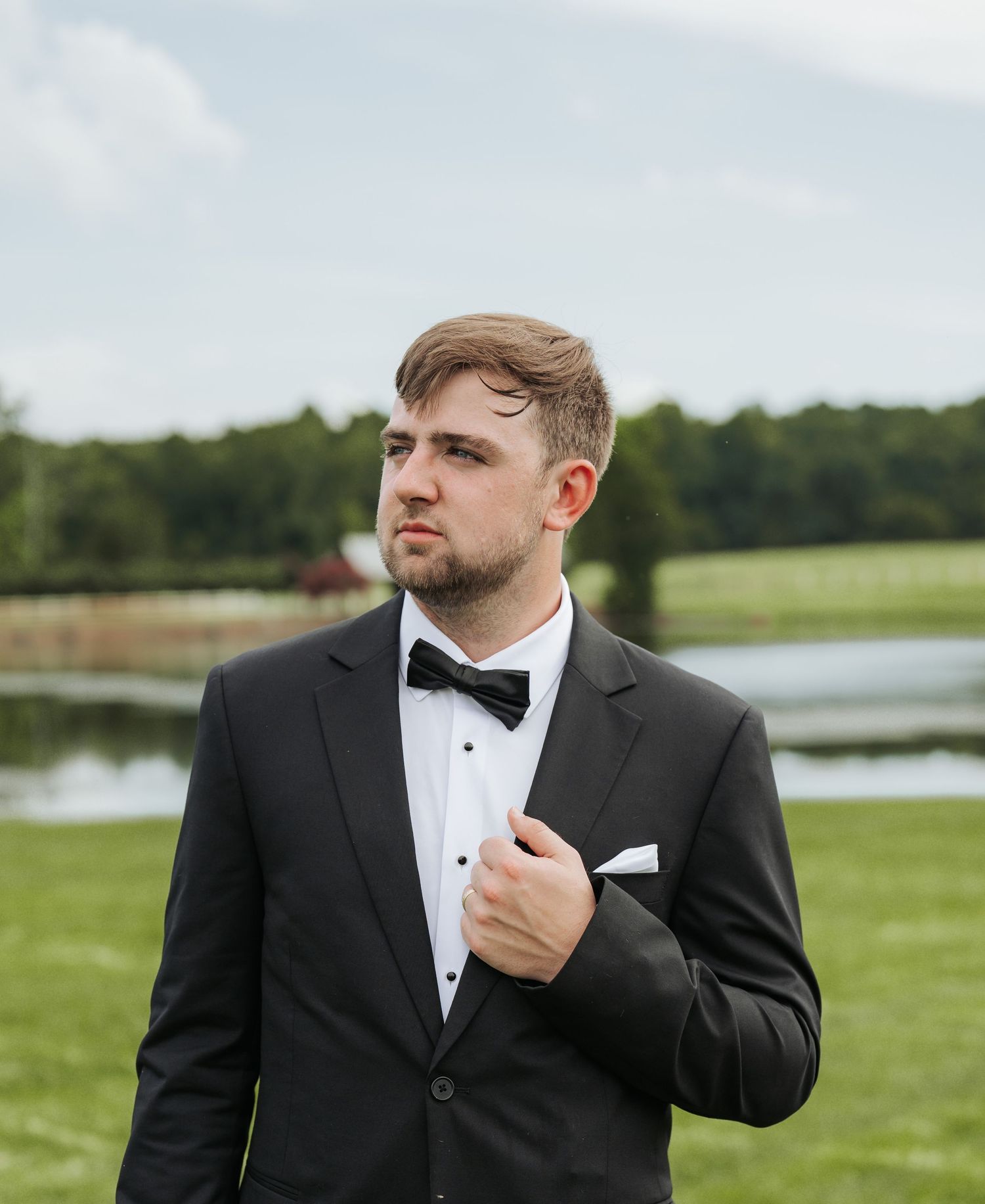 Groom in black tuxedo and bow tie adjusts his collar outdoors on wedding day.