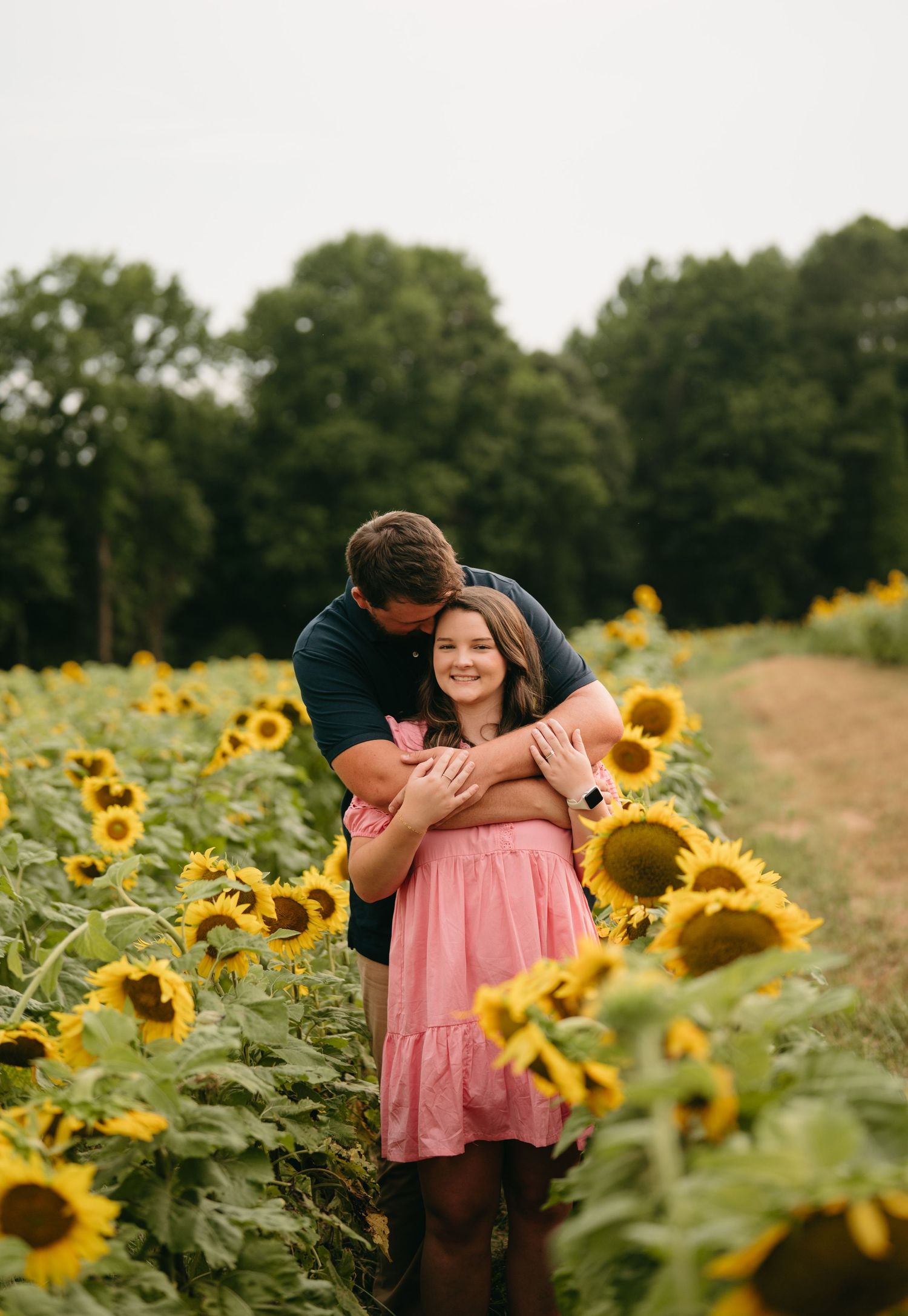 A couple embraces in a golden sunflower field during sunset, creating a romantic summer scene.