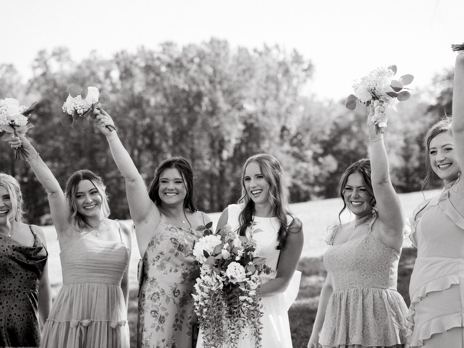 A joyful wedding party celebrates outdoors in black and white photography with trees in the background.