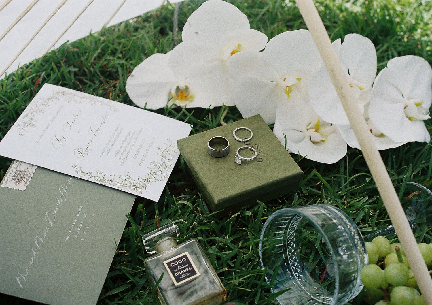 Wedding details with white orchids and invitation cards arranged on grass.