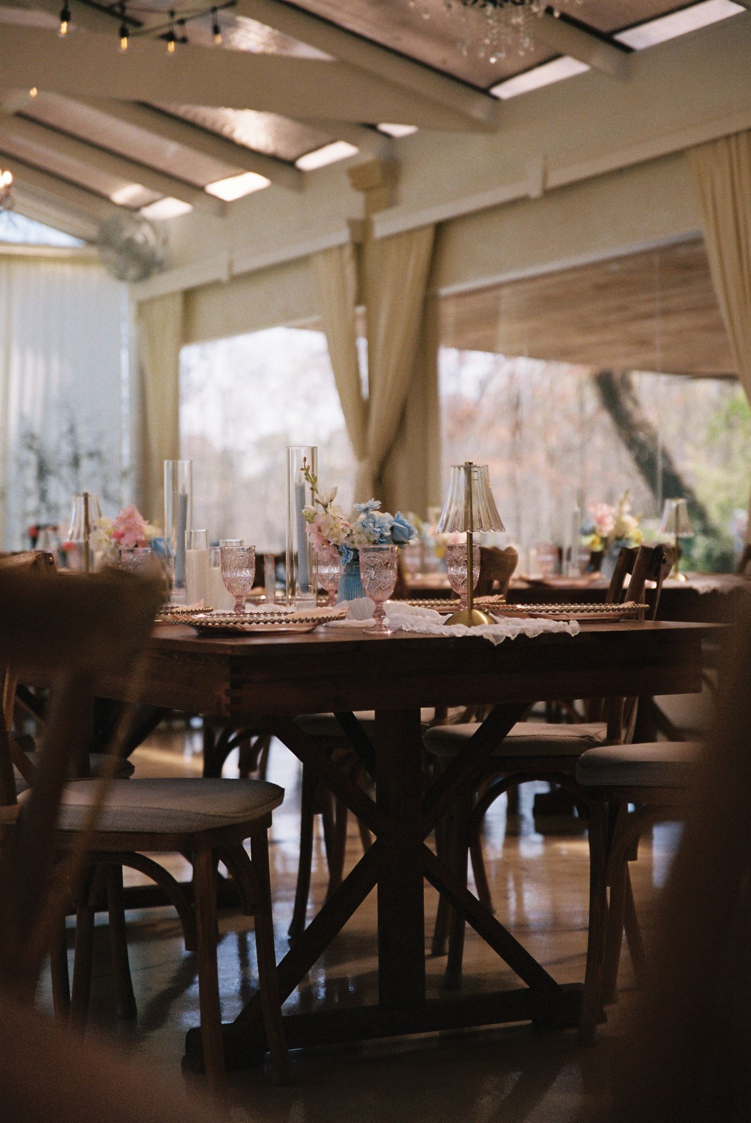 Long wooden dining tables and chairs set up in an elegant event space with soft lighting and sheer curtains.