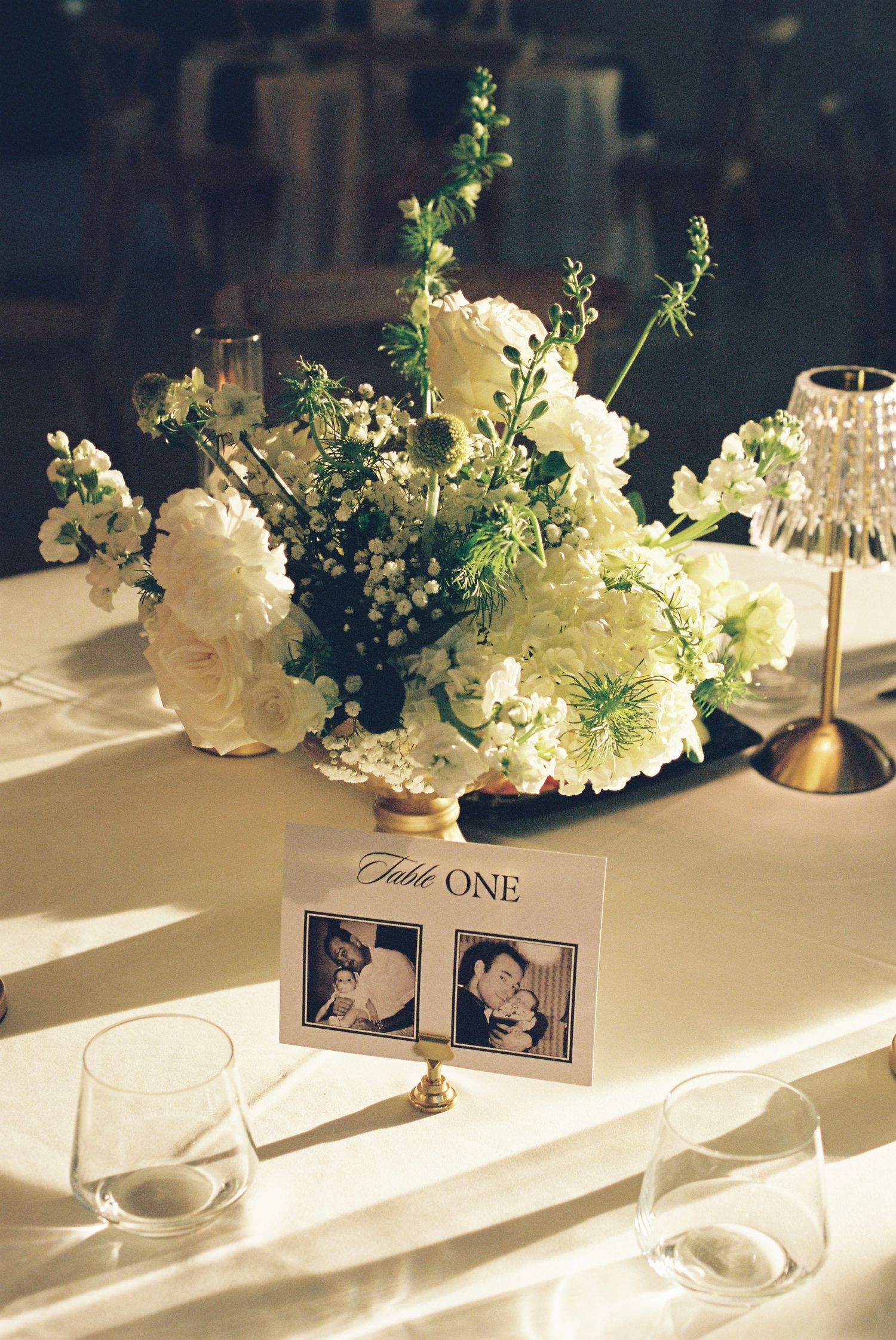 White floral centerpieces and table numbers arranged on elegant wedding reception table captured at golden hour on film.
