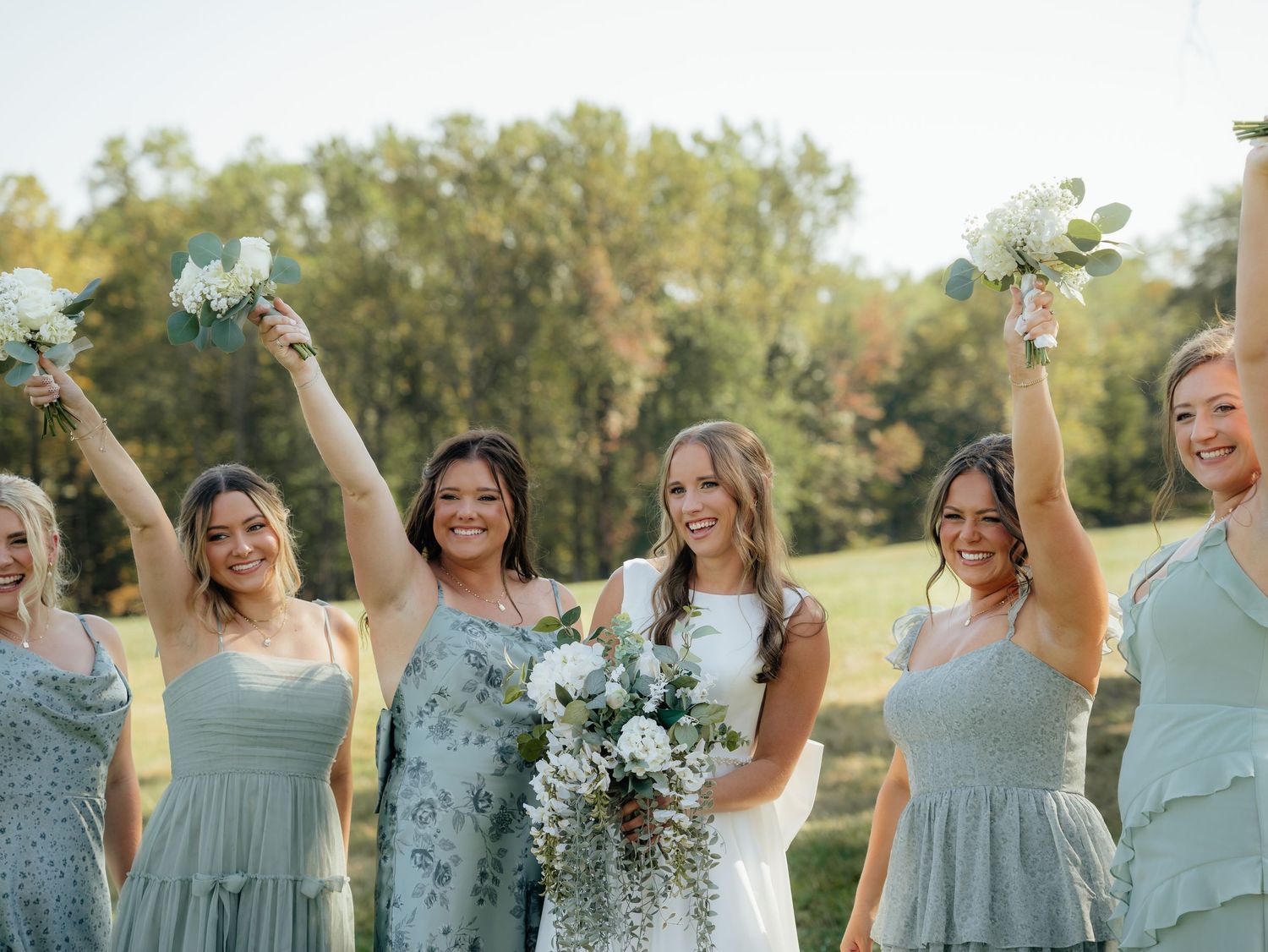 Bridesmaids in sage green dresses celebrate joyfully in a sunny outdoor setting during a wedding celebration.