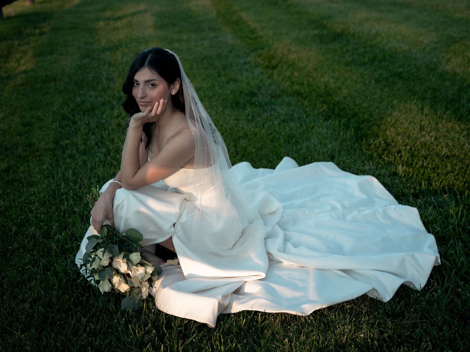 A bride in a flowing white wedding dress lies gracefully on lush green grass while holding a bouquet of flowers.