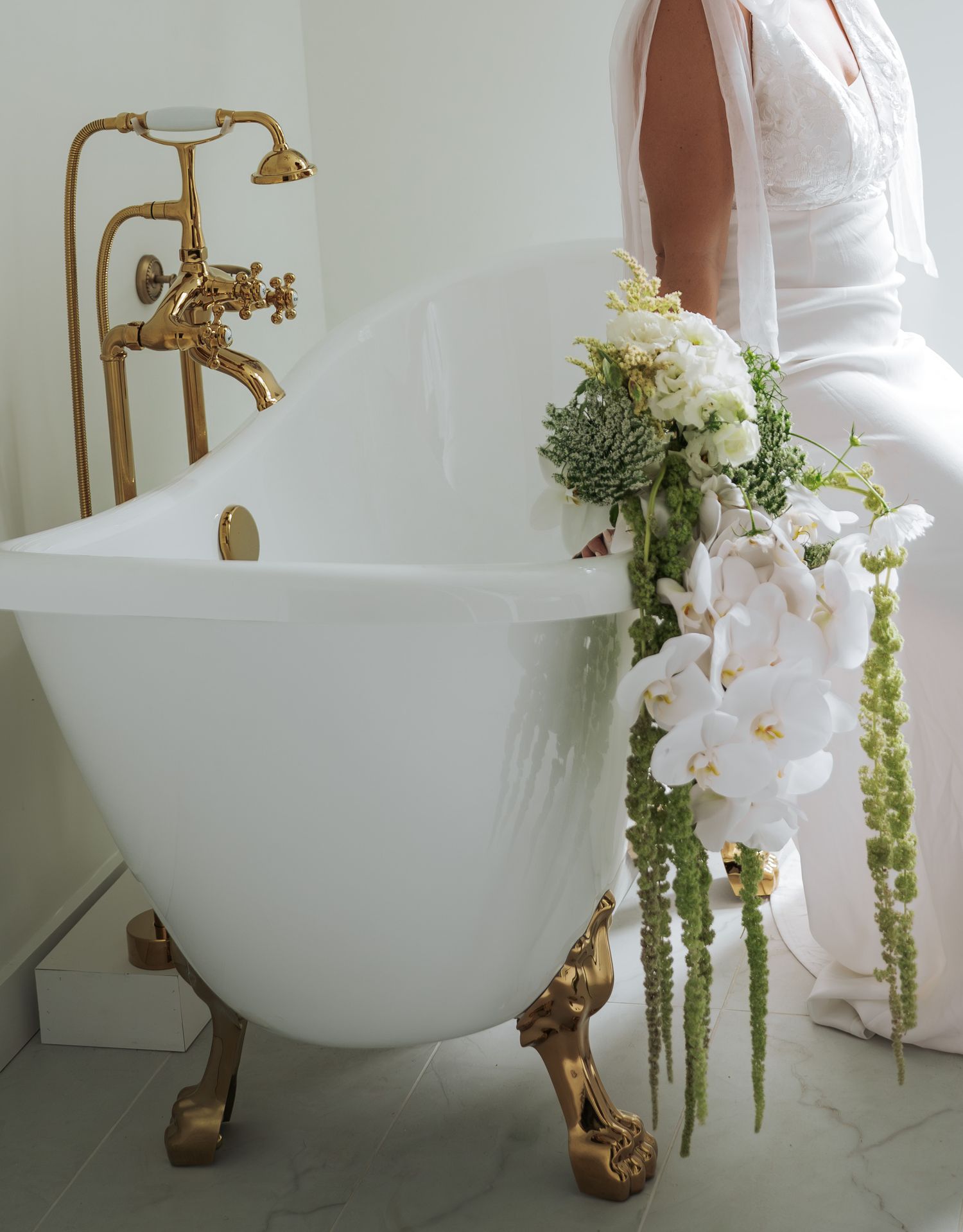 Elegant freestanding bathtub with brass fixtures and cascading greenery arrangement in a minimalist white bathroom.