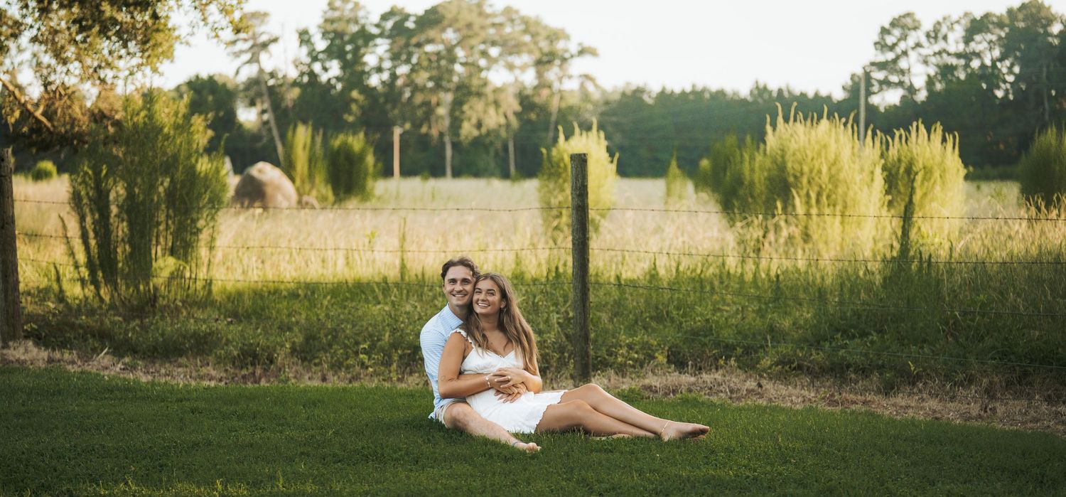 Couple sitting together on green grass in a sunny outdoor field with lush trees and golden light in the background.