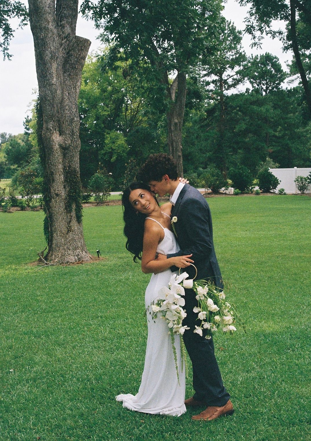 A couple in wedding attire embraces on a scenic green lawn surrounded by mature trees.