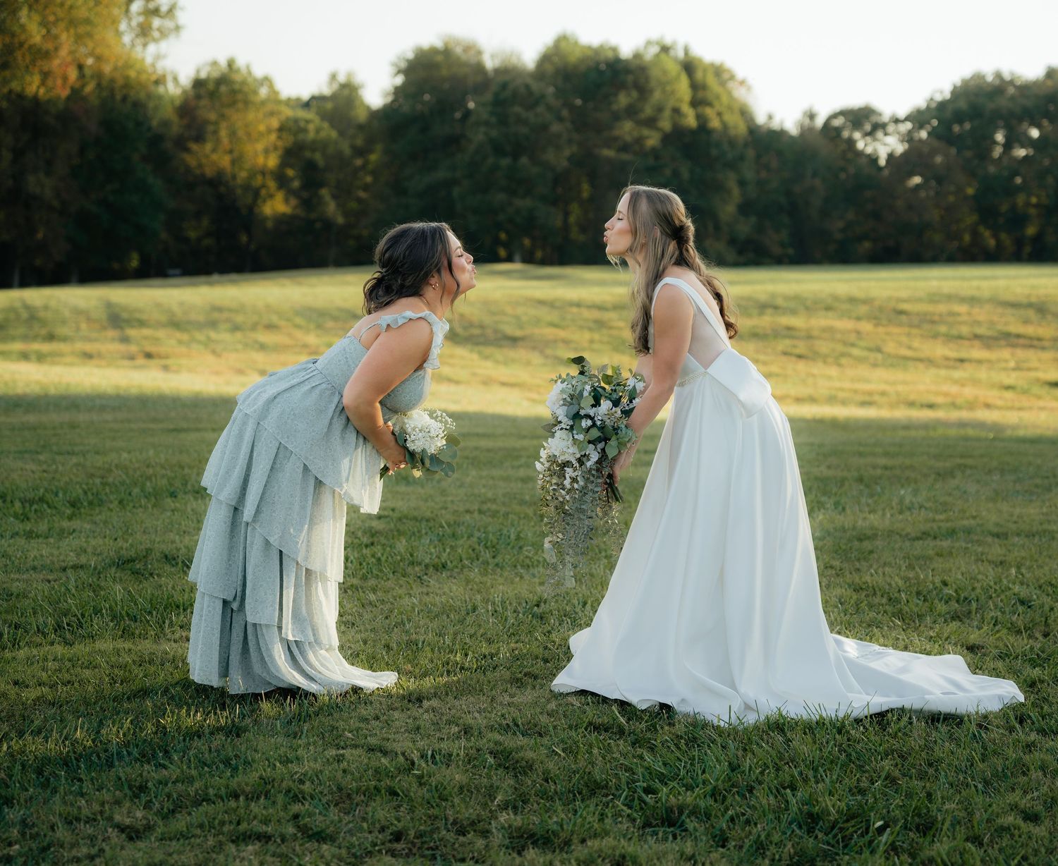 Two people in wedding attire sit together on grassy hillside during golden hour sunset.