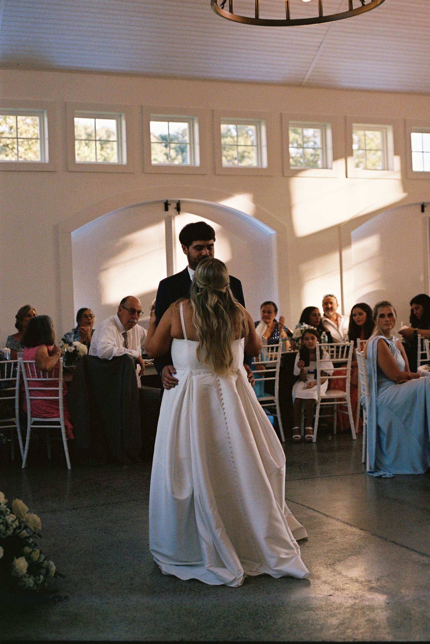 A couple sharing their first dance in an elegant white venue with high windows.