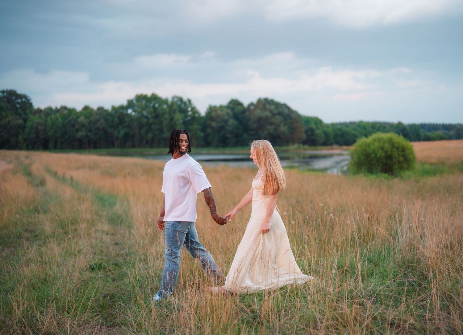Couple holding hands walking through golden meadow field, woman in flowing dress, trees and lake in background.