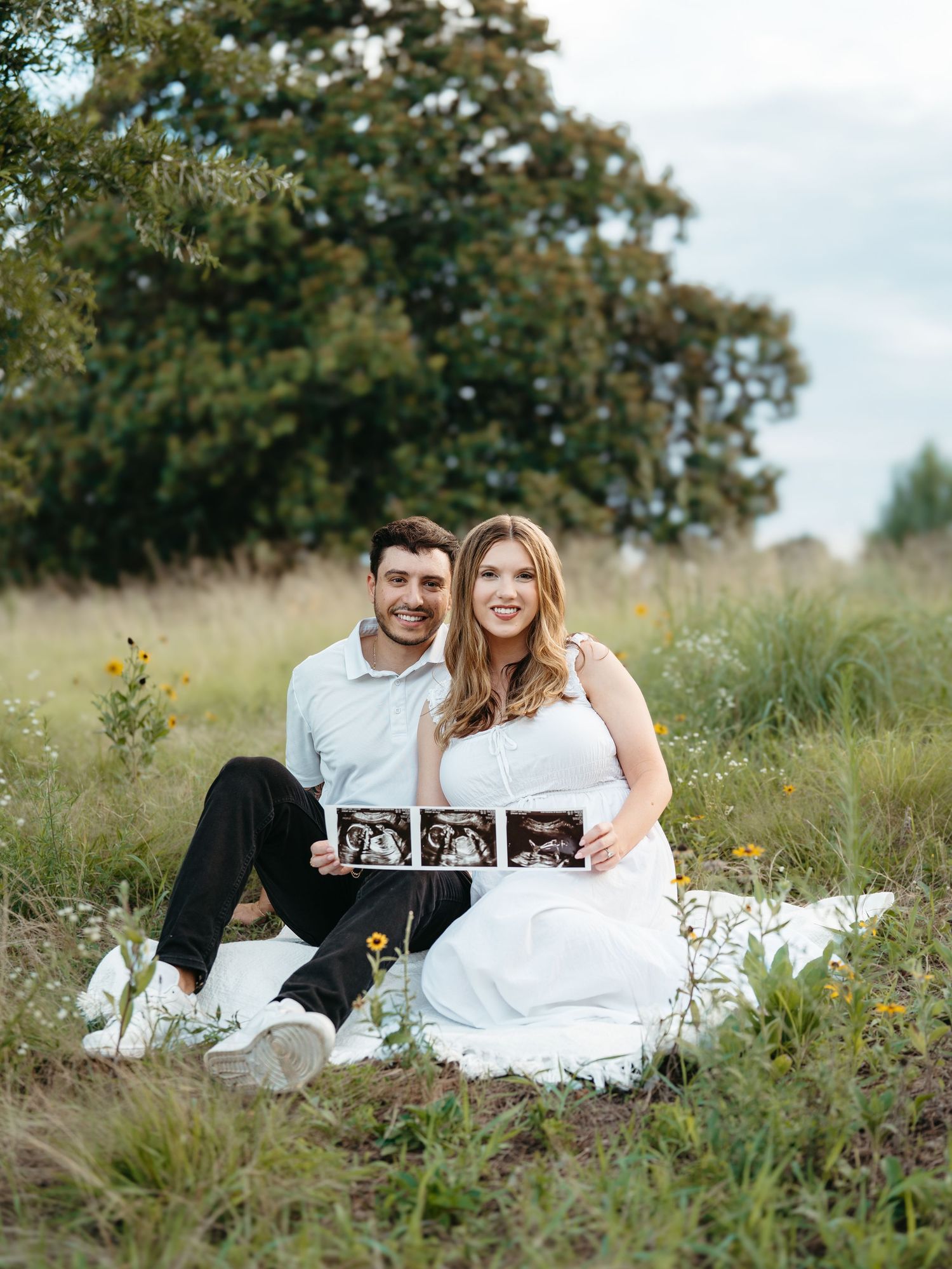 Happy couple sitting in a field holding ultrasound photos during an outdoor maternity announcement photoshoot.