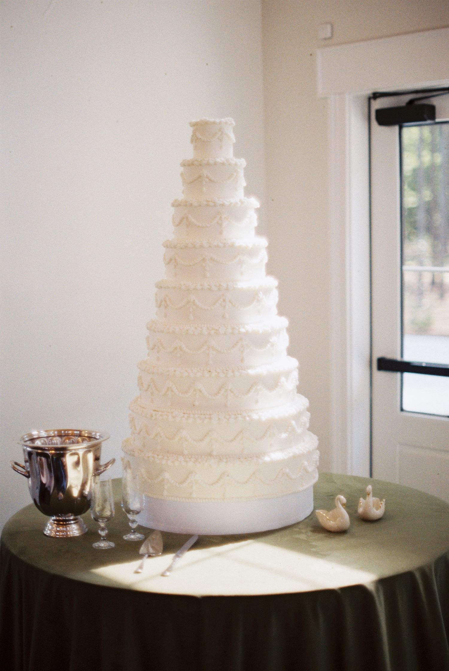 Elegant 10-tier white wedding cake with intricate piped lace details on a green draped table near a window.