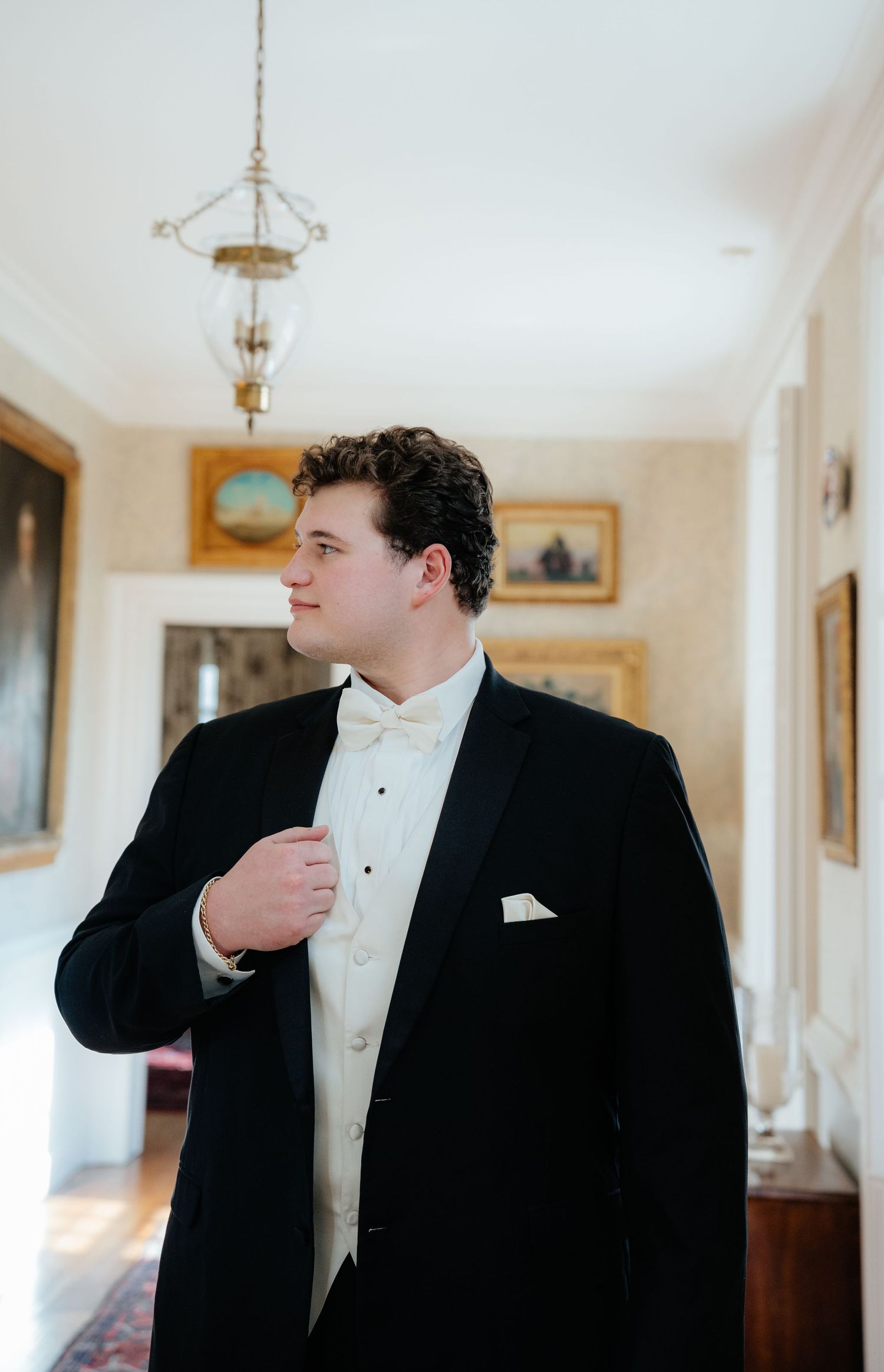 A person in formal black tuxedo and white dress shirt stands in an elegant hallway with white walls and decor.