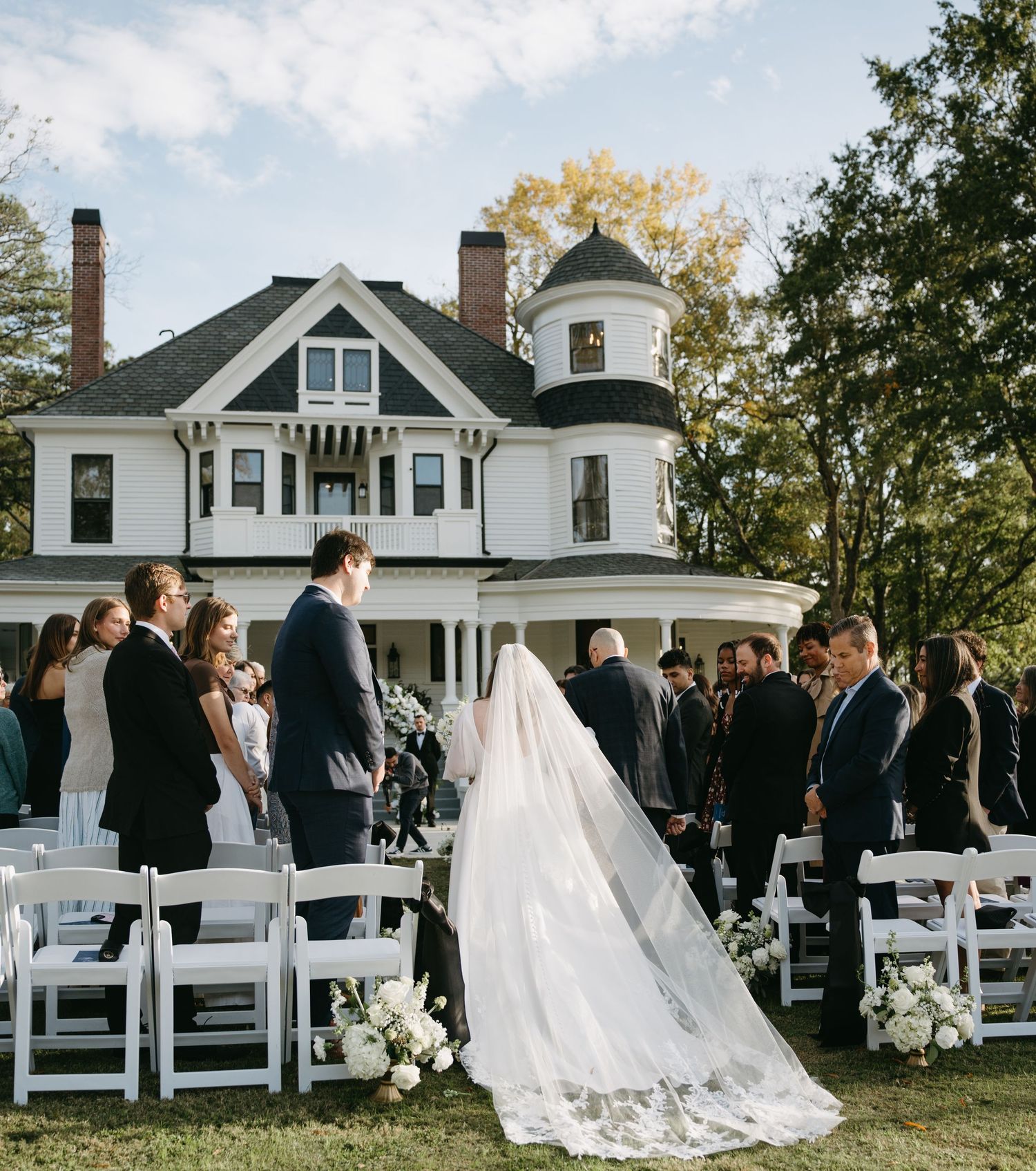 Outdoor wedding ceremony setup with white chairs facing a historic white farmhouse in an autumn setting.