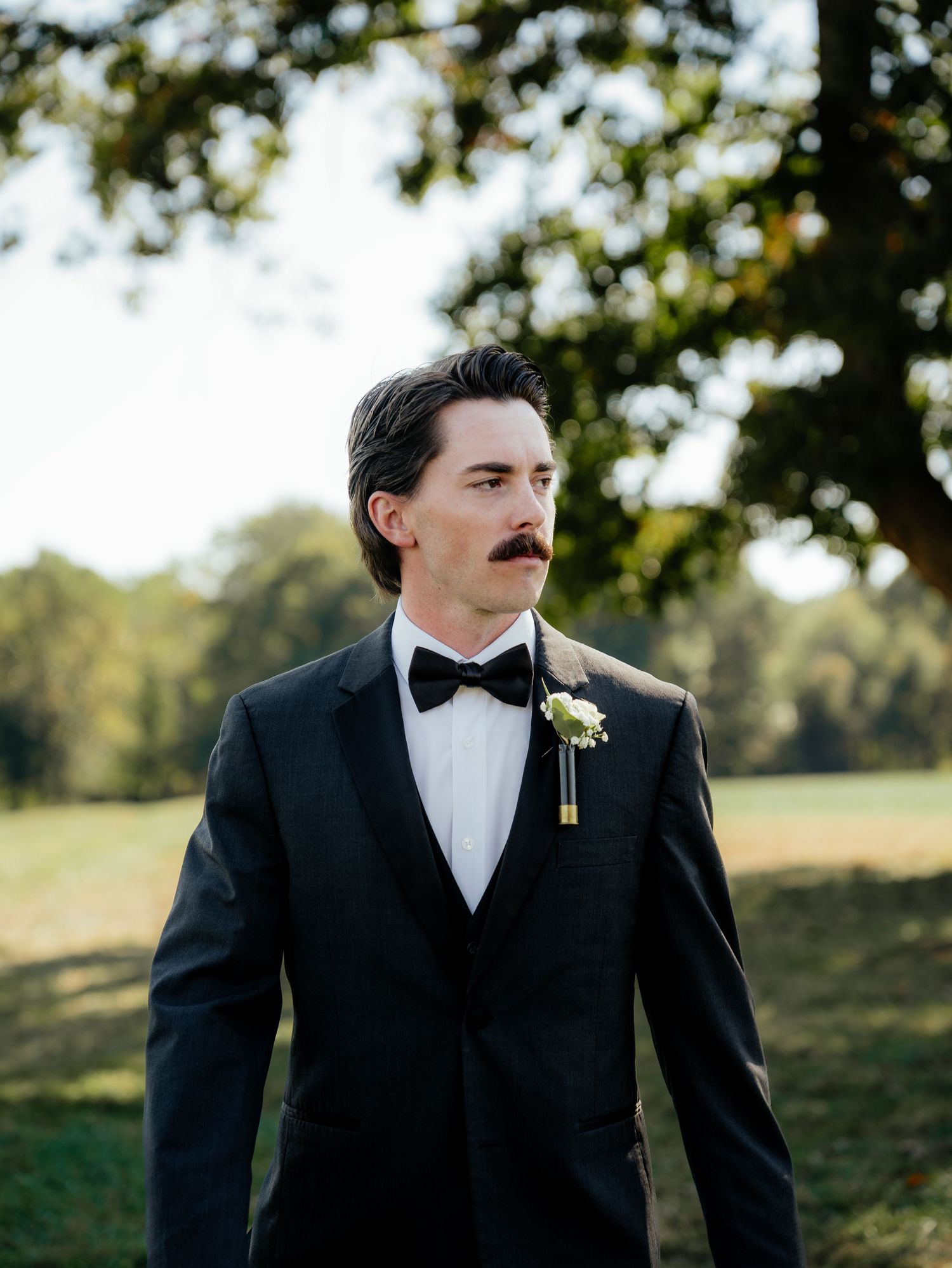 Groom in classic black tuxedo and bowtie poses outdoors at sunset.
