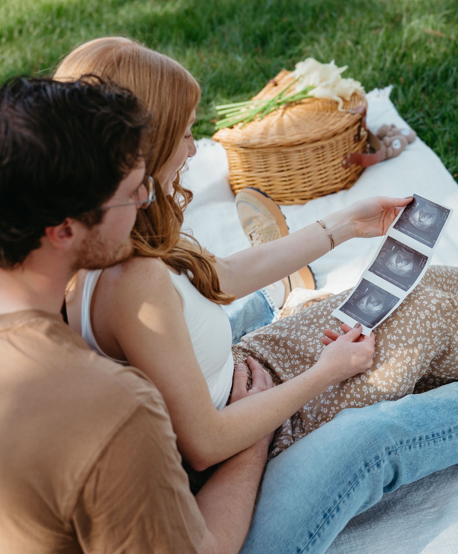Couple sitting on a picnic blanket outdoors, looking at ultrasound photos together near a wicker basket.