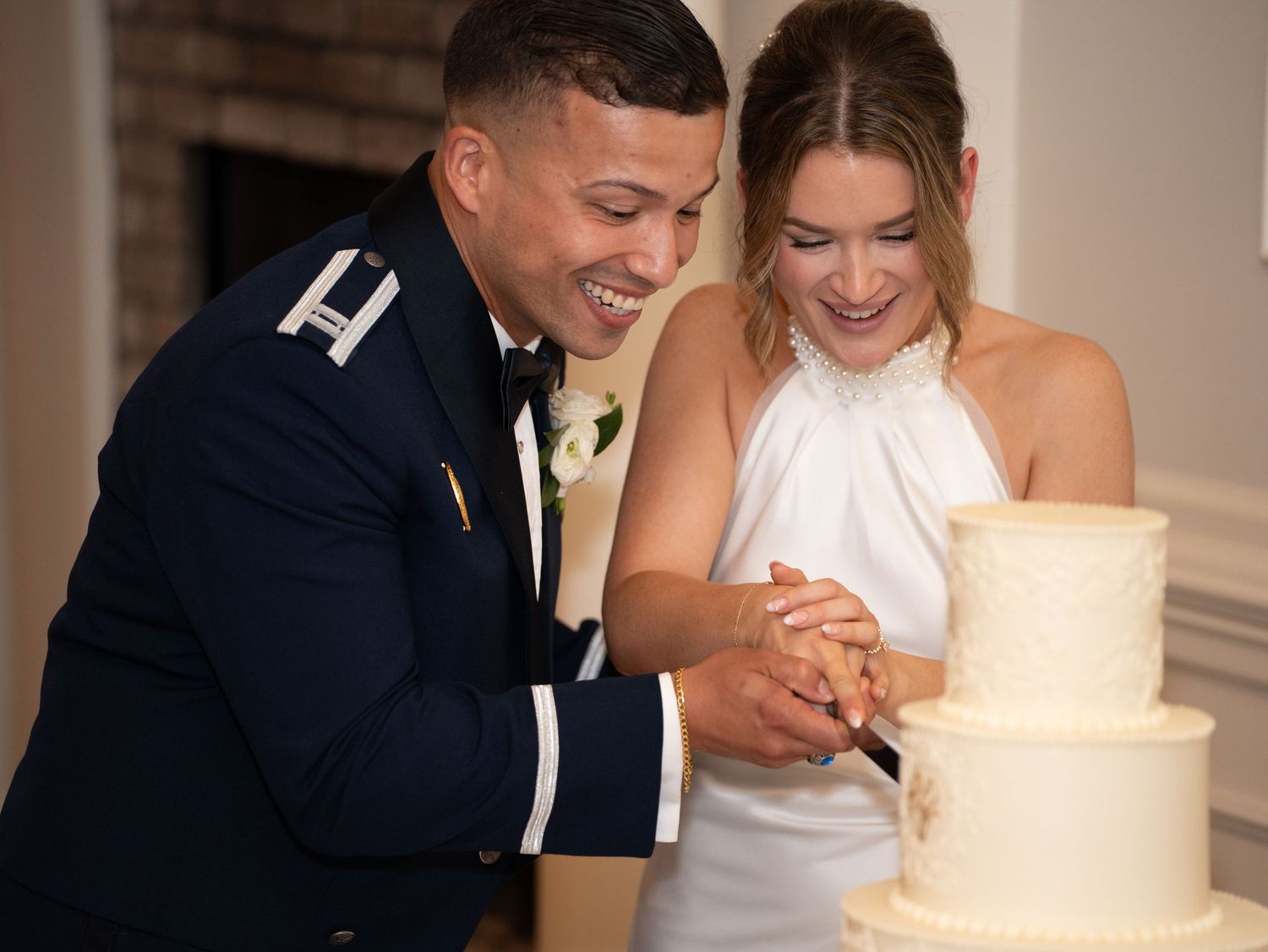 Military groom and bride in white halter dress cutting wedding cake together, smiling joyfully at elegant reception.