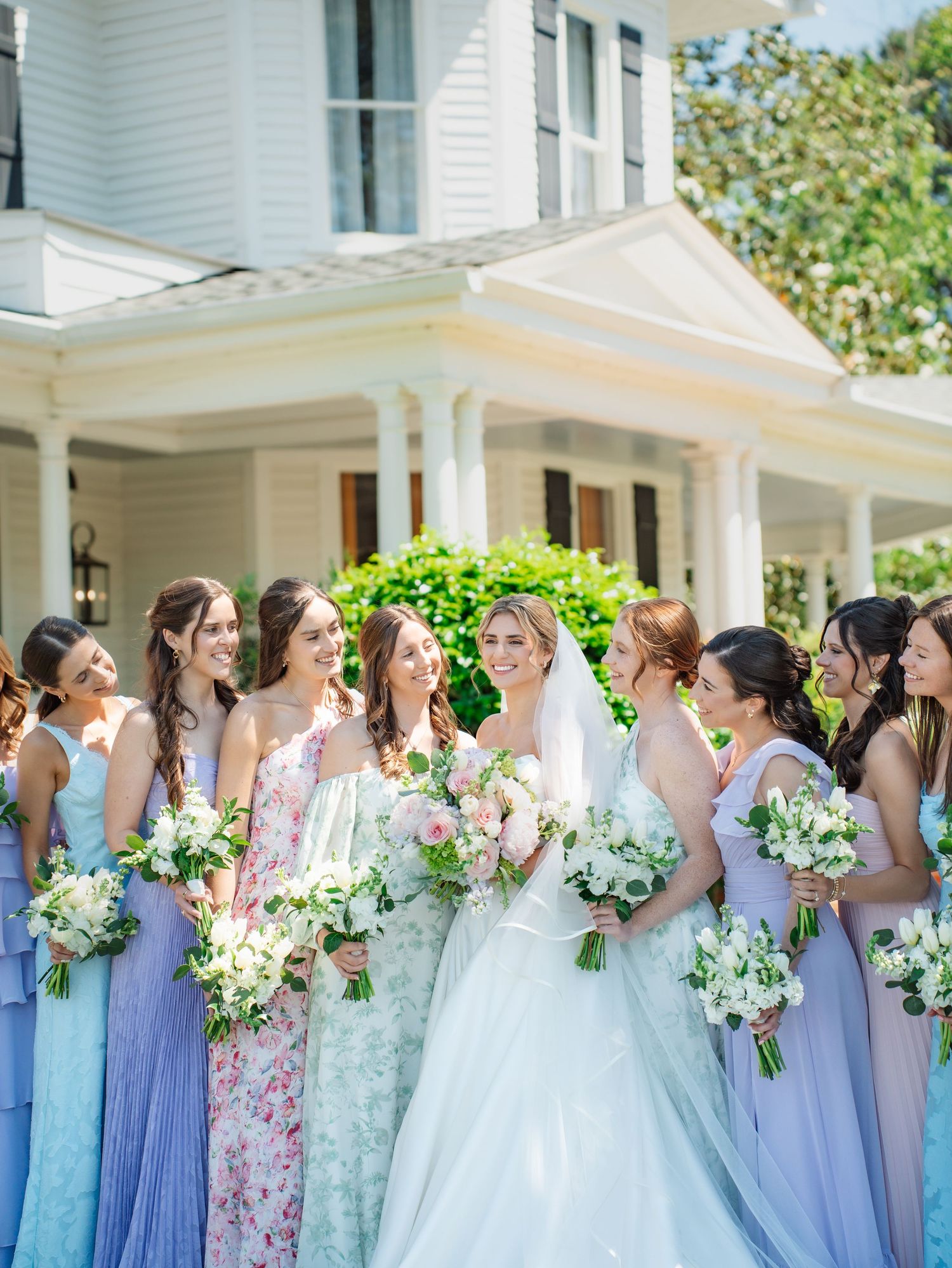 Bride in white gown with bridesmaids in pastel dresses holding bouquets outside elegant white mansion.