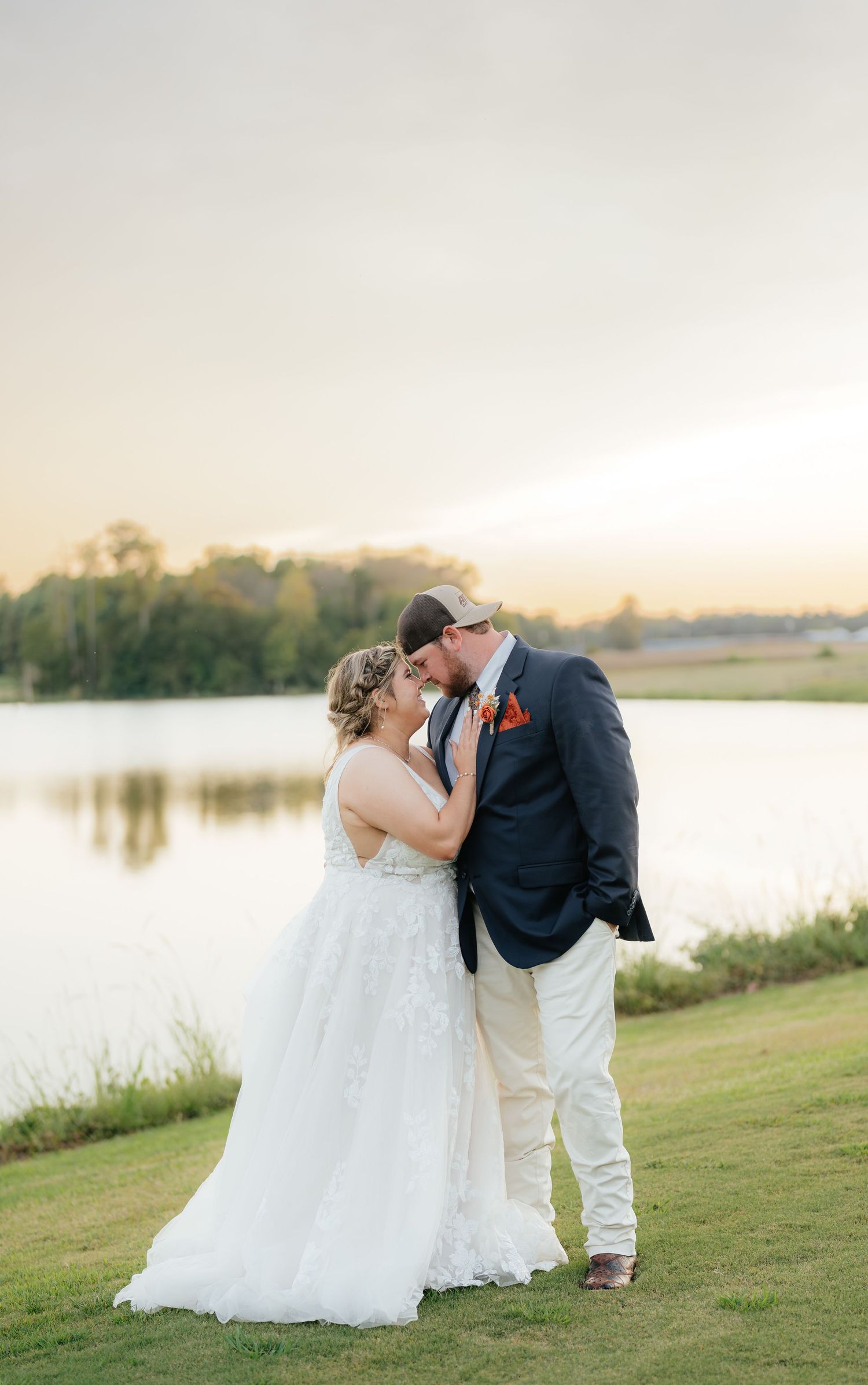 A romantic sunset wedding moment captured by a lake with a couple embracing on a grassy shore.