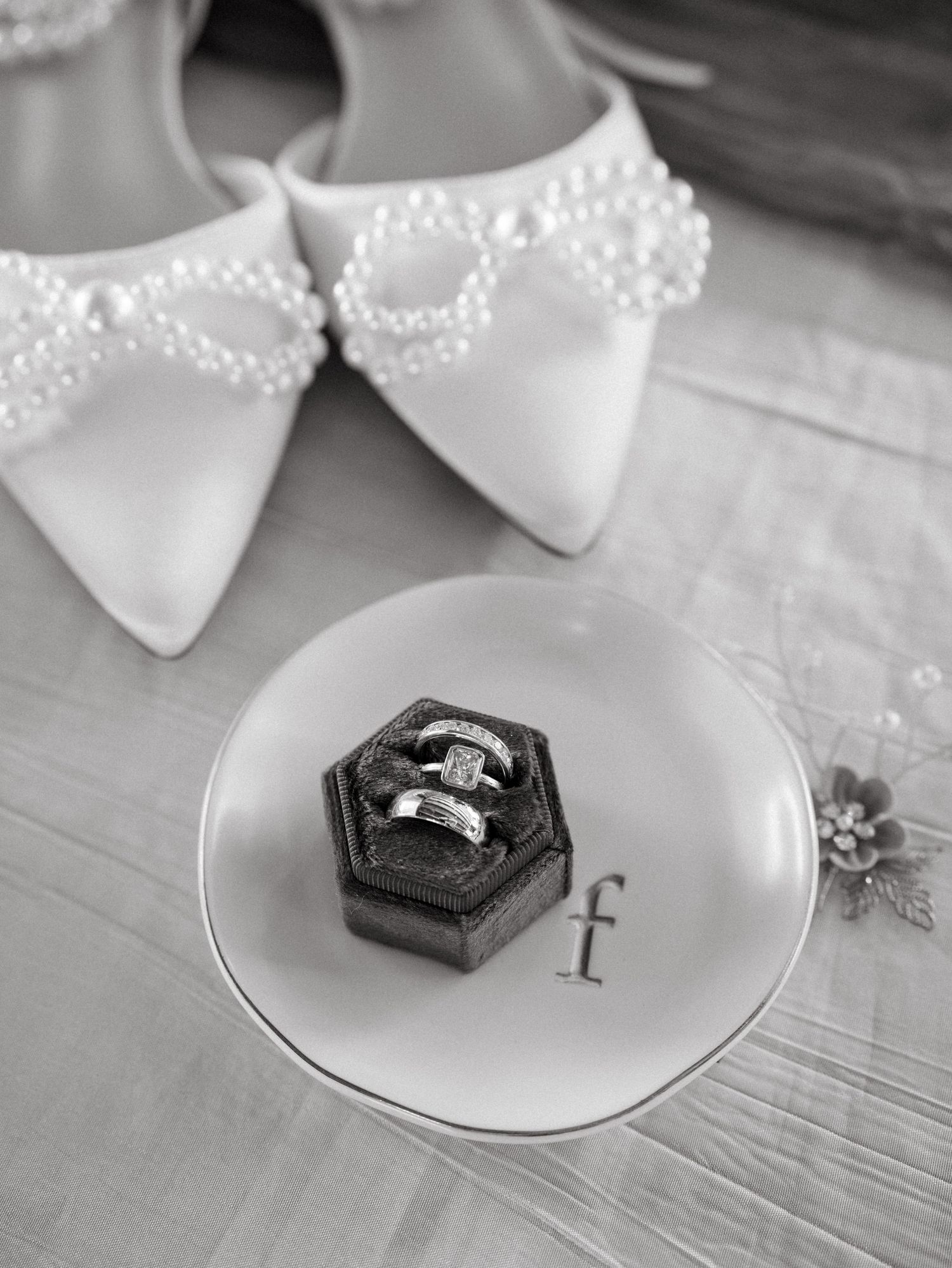 Wedding rings displayed in a black hexagonal box on a white plate with elegant white shoes in the background.