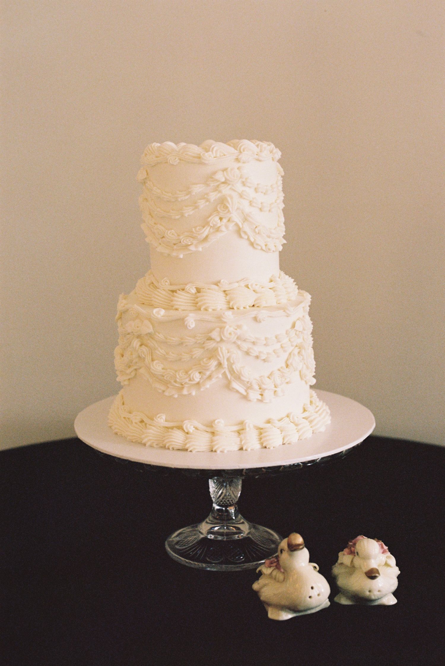Elegant white wedding cake with delicate floral details displayed on a glass stand.