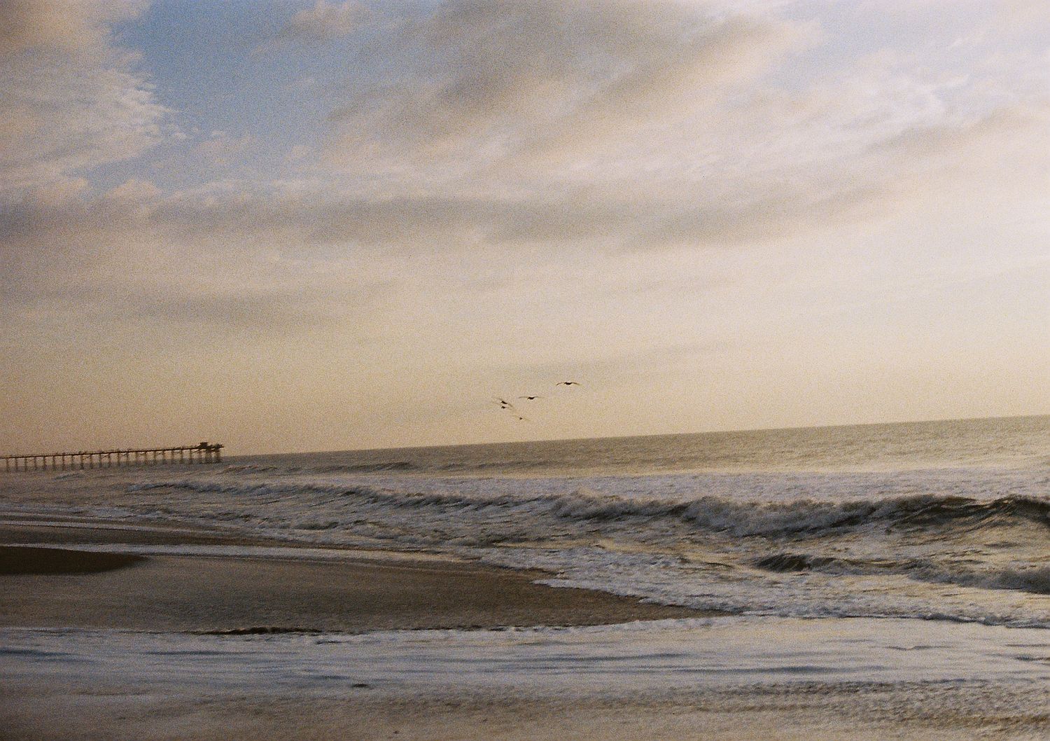Dramatic waves crash along a beach shoreline during golden hour with a pier in the distance.