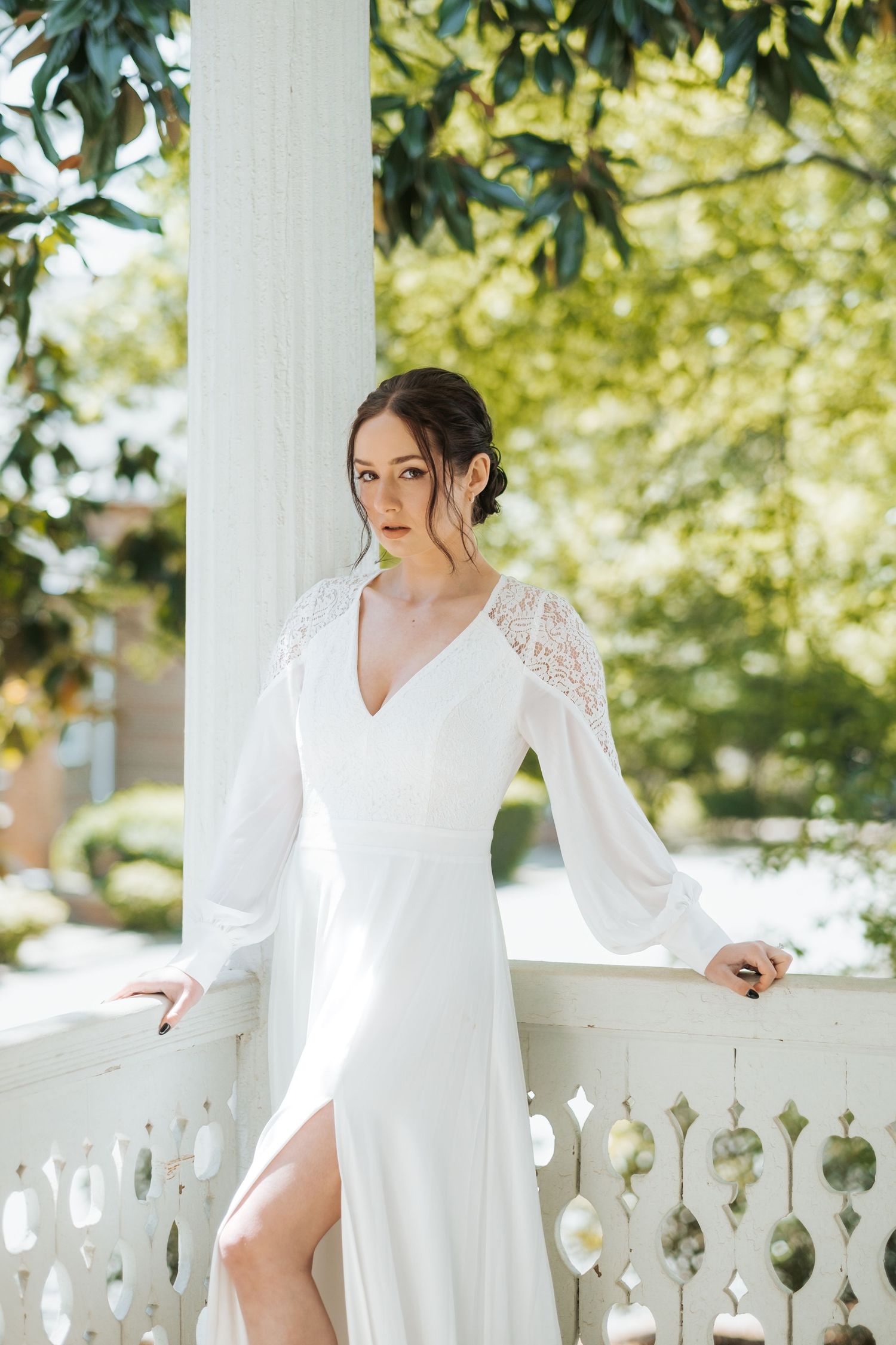 A woman in a white lace dress poses elegantly on a classic white porch balustrade in a garden setting.