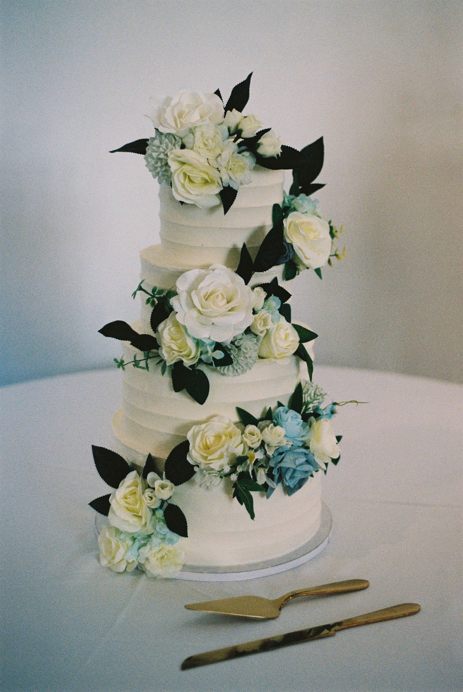 Three-tiered white wedding cake decorated with cream roses and green foliage against light background.
