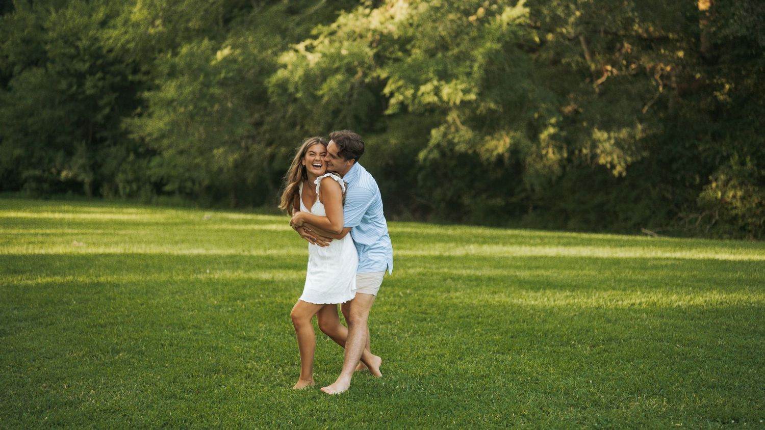 A couple embraces and shares a kiss on a lush green lawn surrounded by trees in a sunny park.