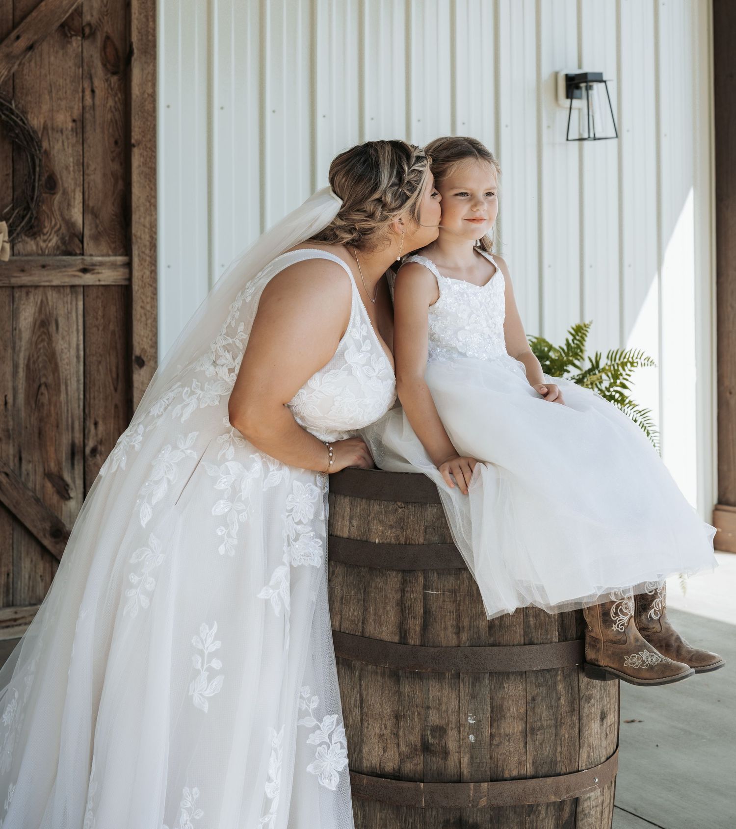 A tender moment shared between a bride and young attendant sitting on a rustic wooden barrel at a barn venue.
