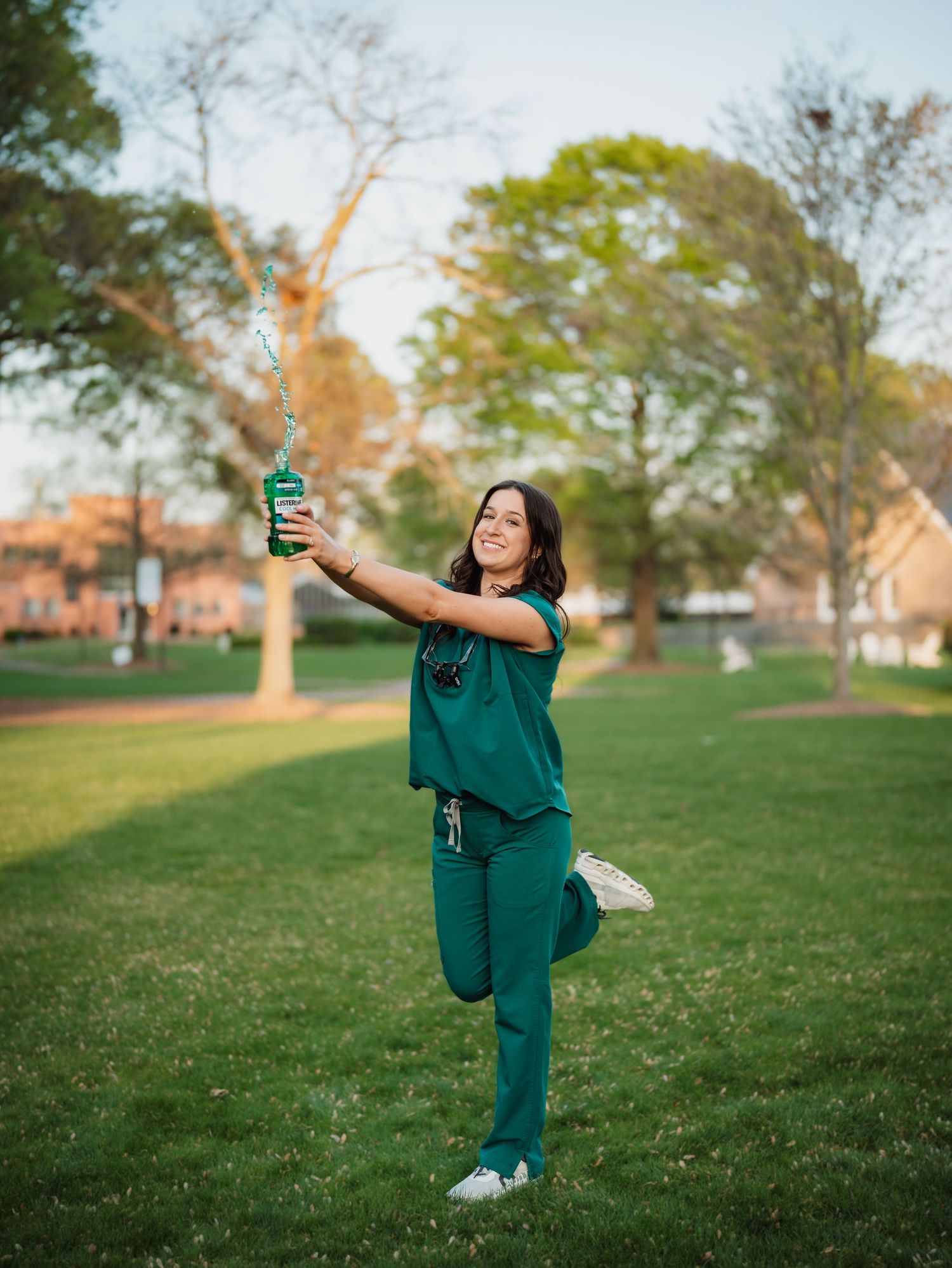 A nurse in green scrubs poses playfully in a park, balancing on one leg while holding a green bottle aloft.