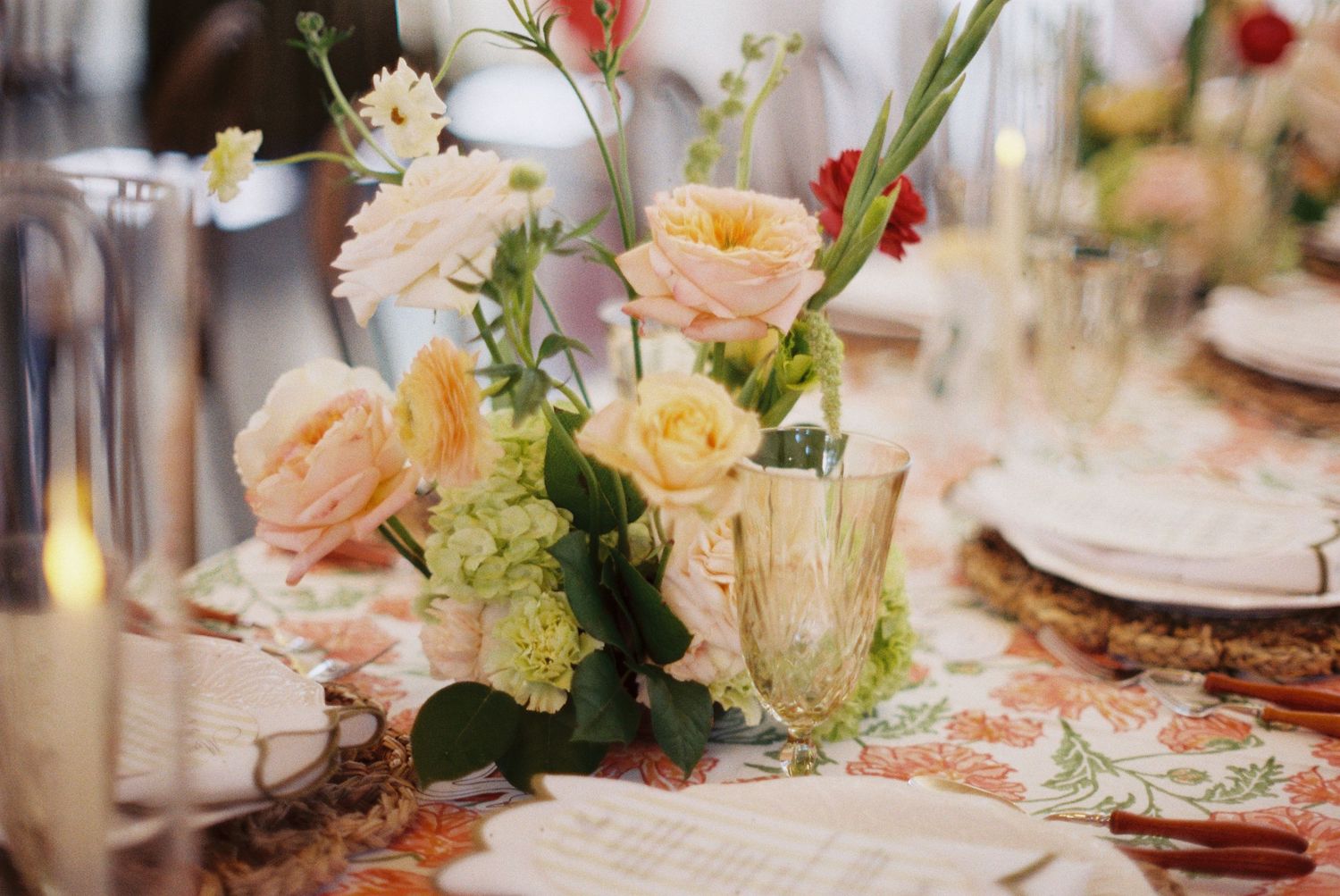 Elegant wedding table centerpiece with peach roses, green hydrangeas, and red accents on a floral patterned tablecloth.