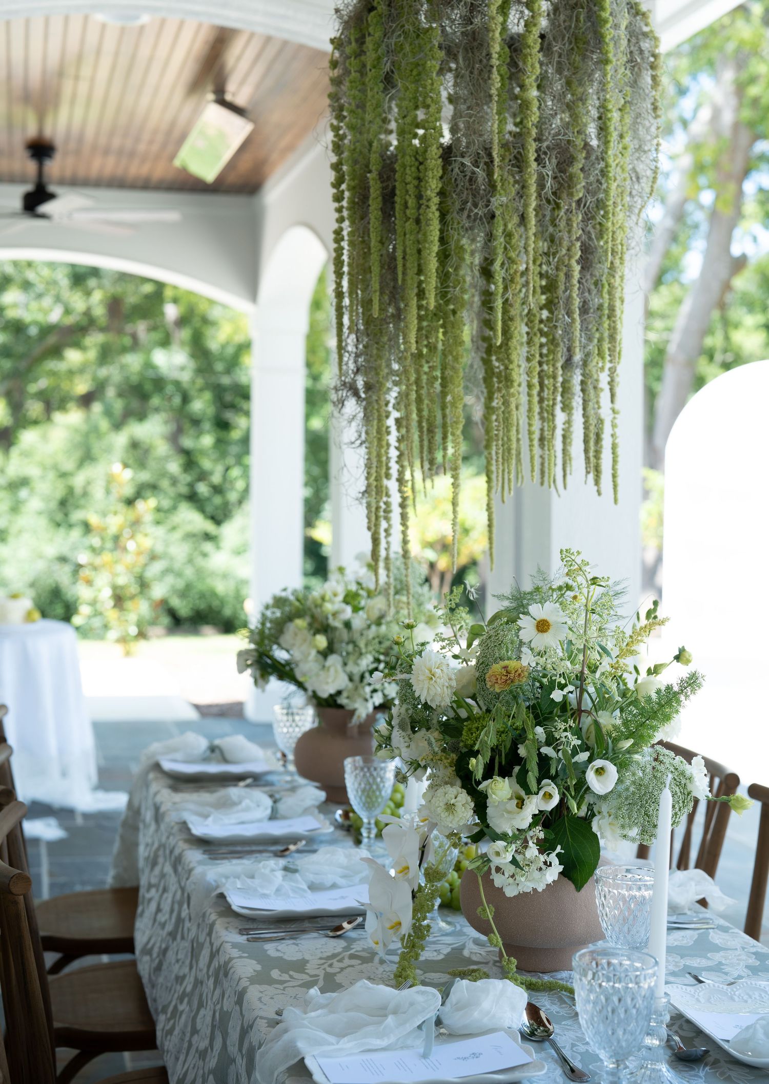 Elegant wedding reception table settings with hanging amaranthus and white floral centerpieces under a covered porch.