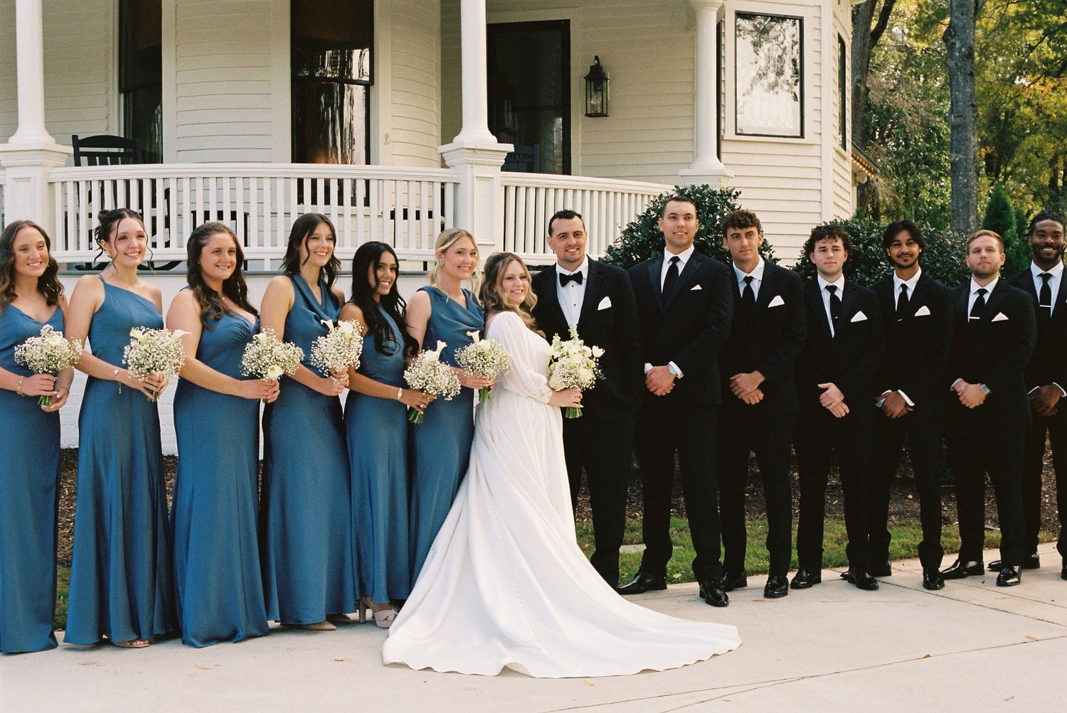 Wedding party in blue dresses and black suits lined up on a classic white porch.