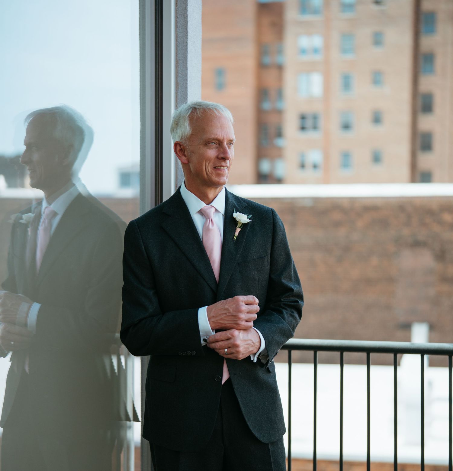 Elegant man in dark suit and pink tie stands by large window with city building reflection.