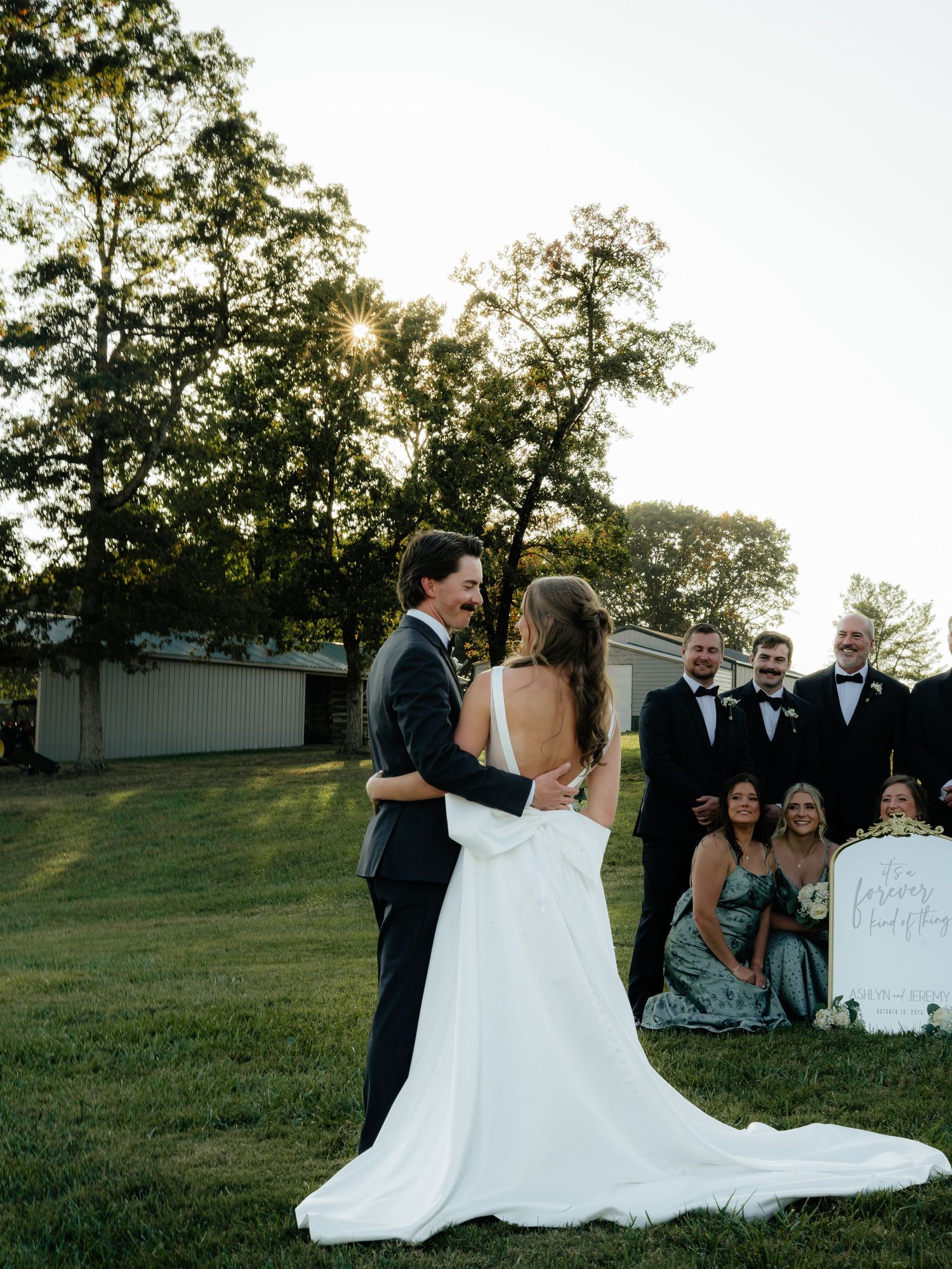 Wedding party gathers on a grassy hillside at sunset for outdoor ceremony and group photos.