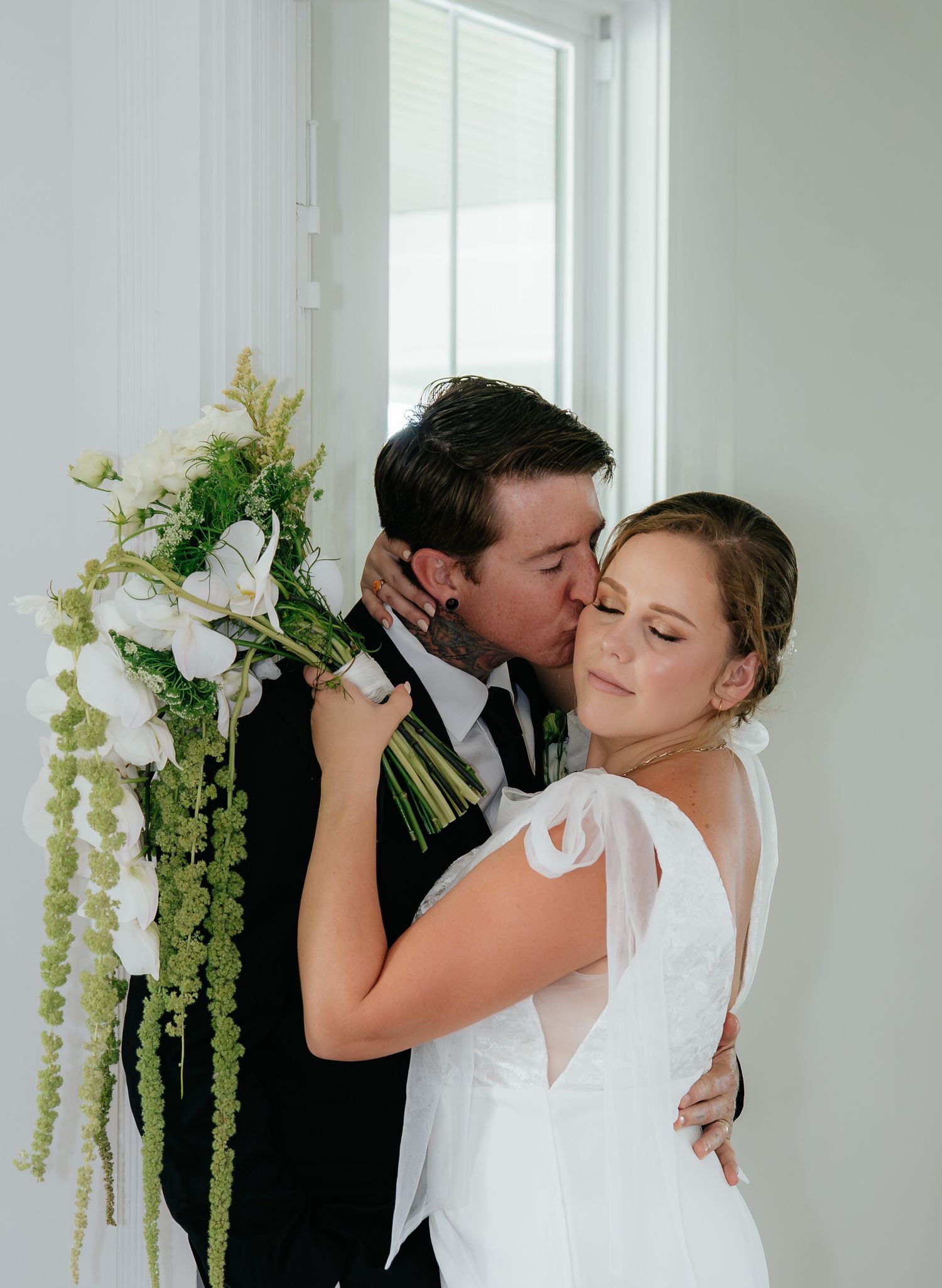 A bride and groom share a romantic kiss in a bright white room decorated with hanging green floral arrangements.