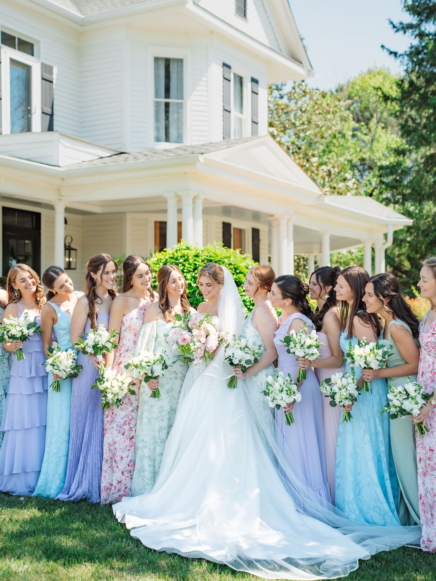 Bride in white gown with bridesmaids in pastel purple and floral dresses holding bouquets outside a white Victorian home.