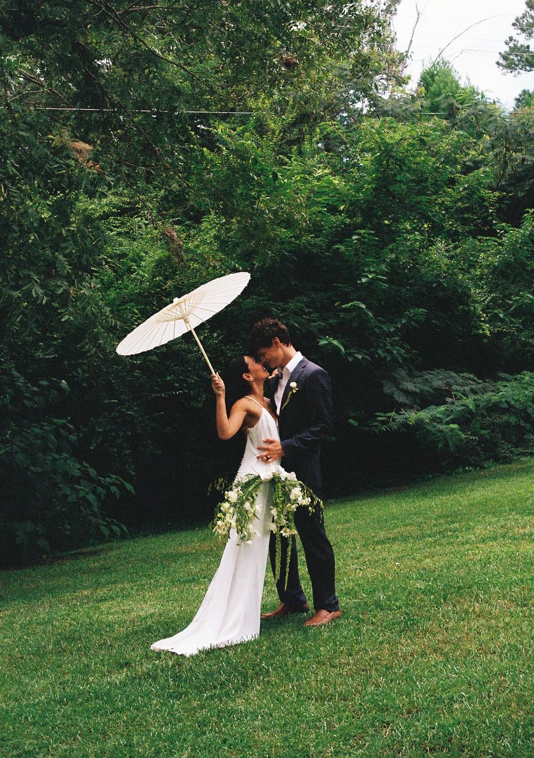 Couple poses romantically with white parasols in a garden setting.